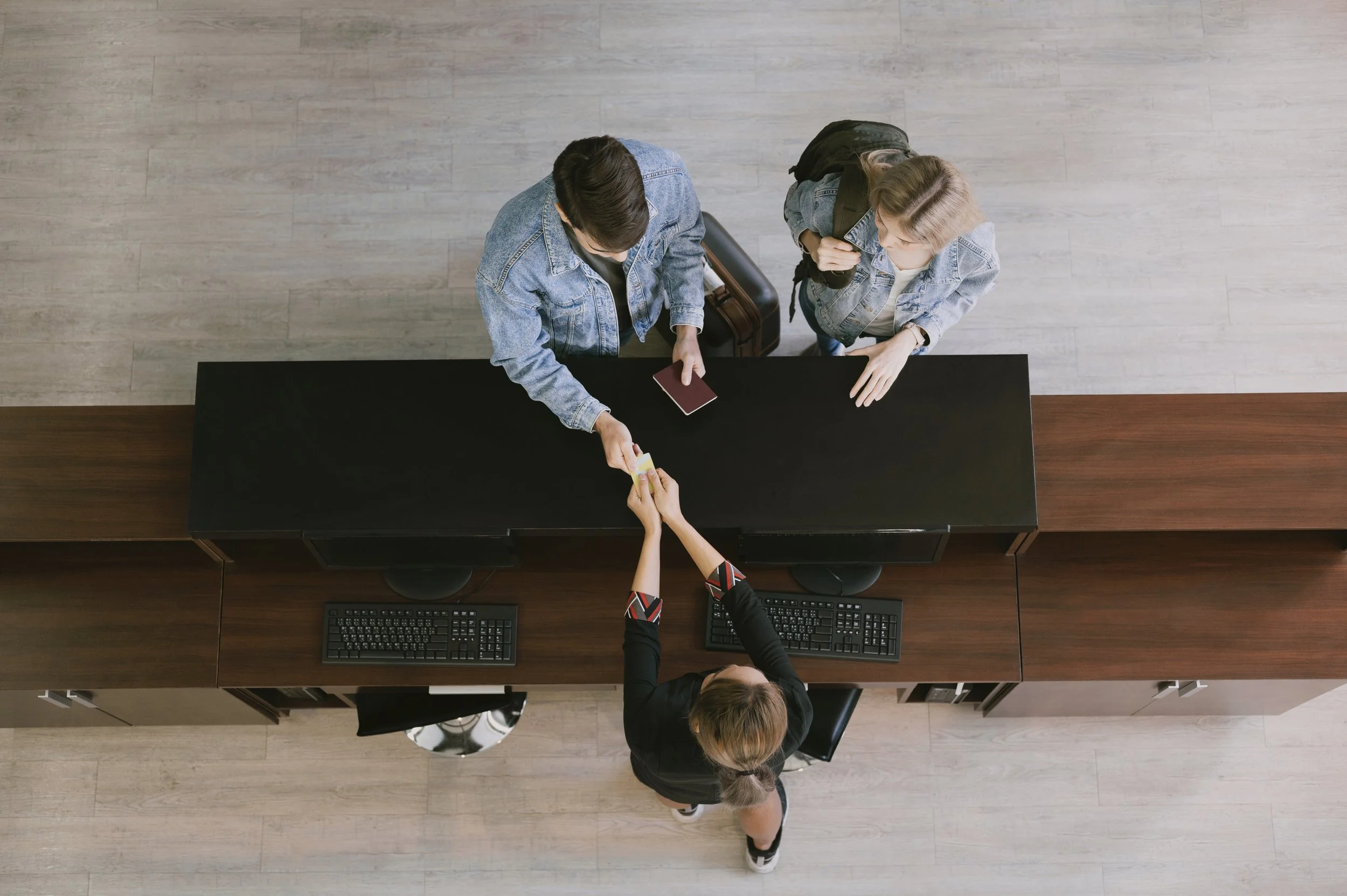 Two women at a reception desk, one handing over a card or document to a staff member, in an office or lobby setting.