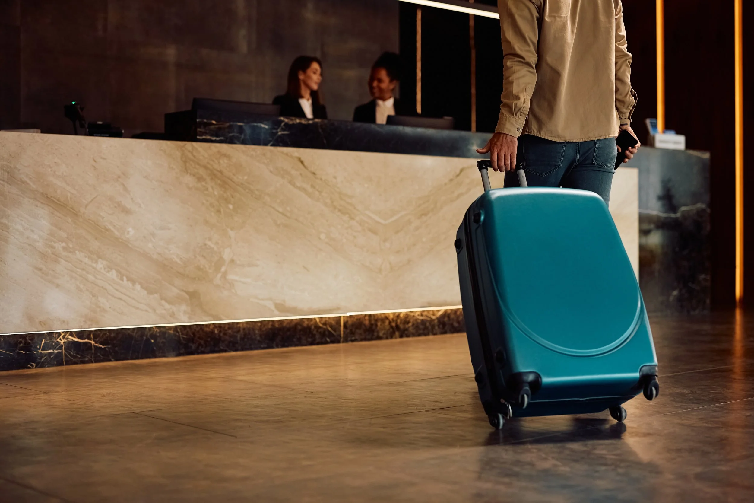 A person holding a suitcase and a smartphone waiting at a hotel reception counter with two smiling receptionists.