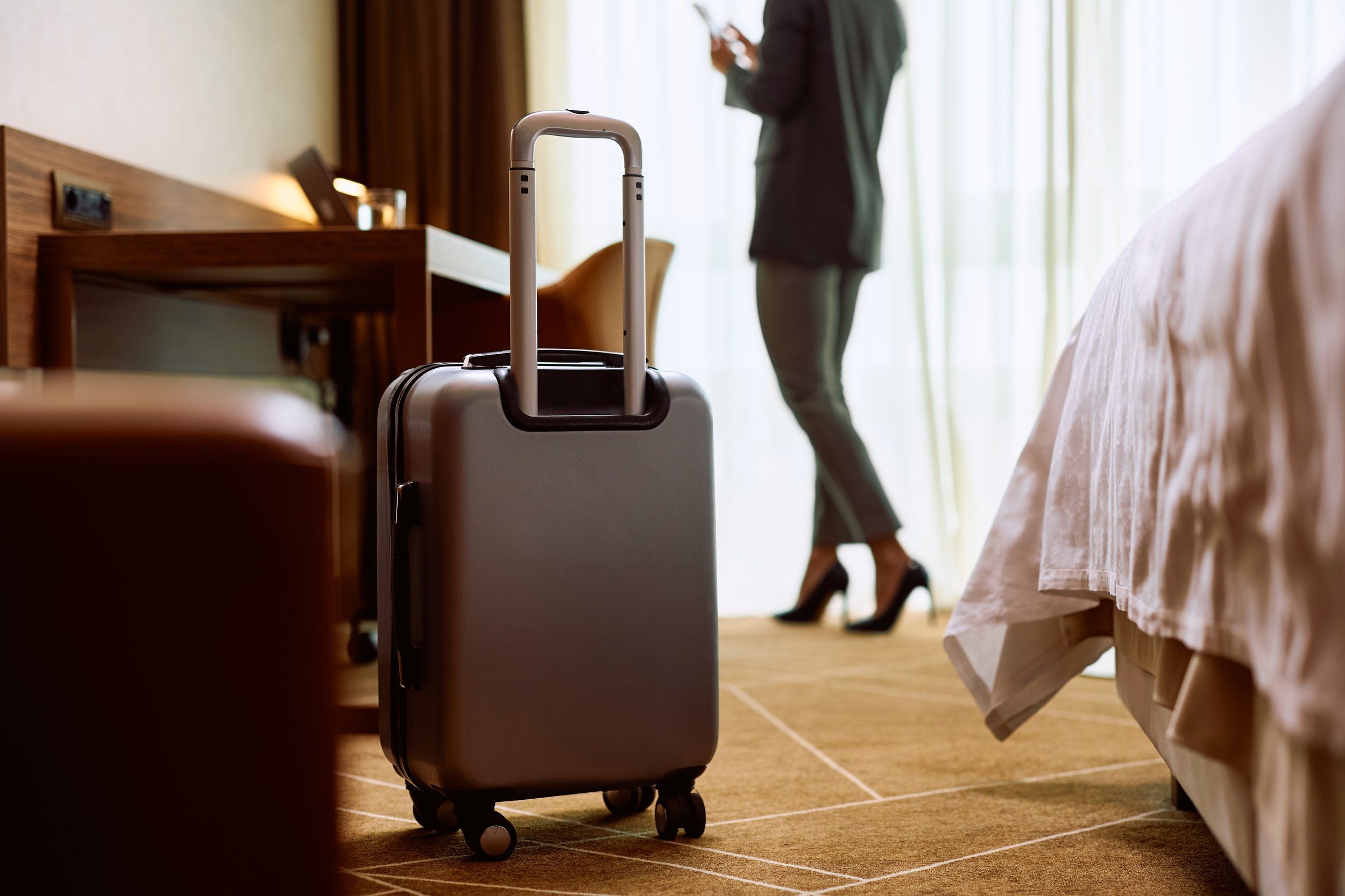A silver rolling suitcase standing on a wooden floor near a bed, with a person in business attire standing in the background by a window with sheer curtains.