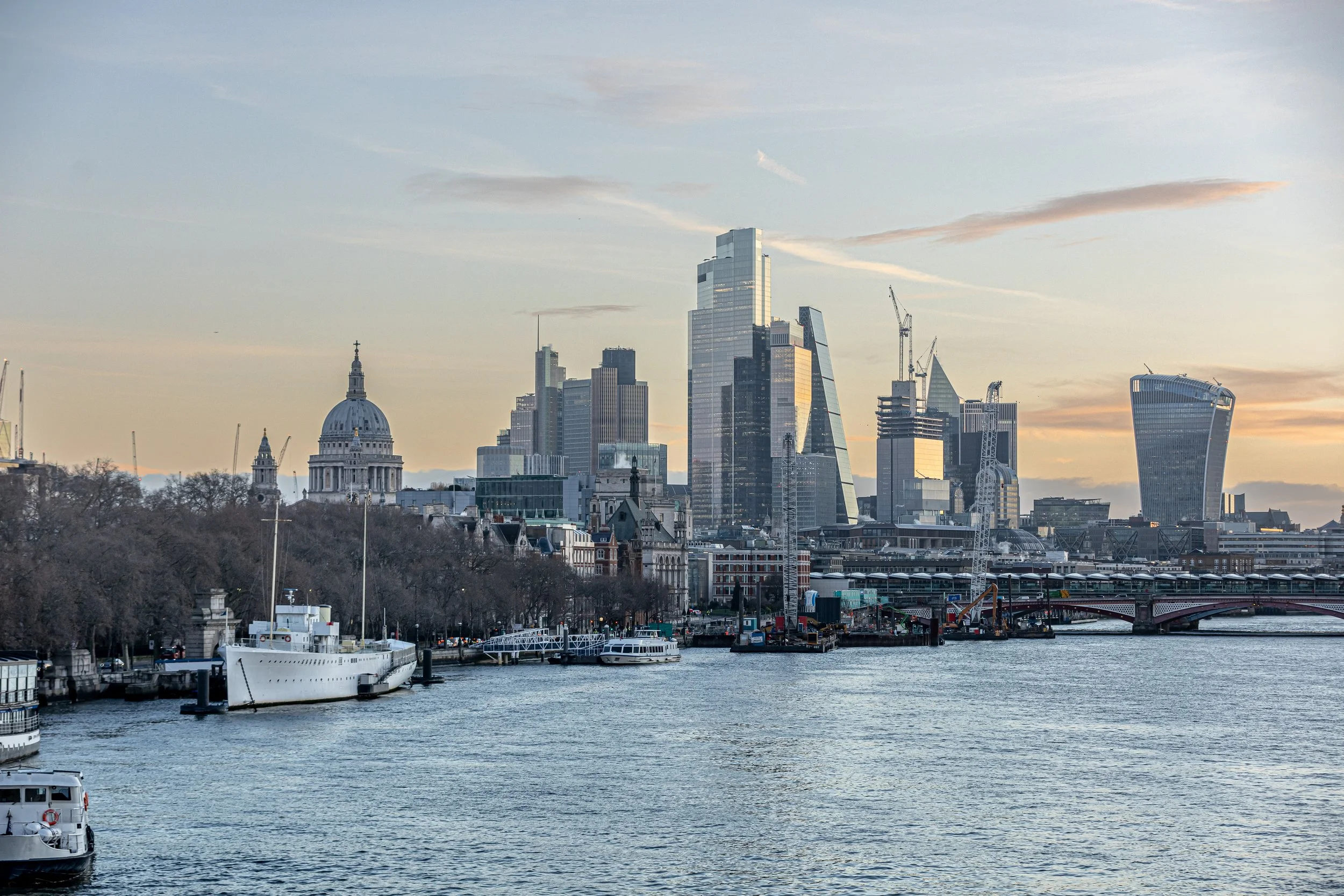 View of London skyline along the River Thames during sunset, with modern skyscrapers and historic buildings, boats docked along the river.