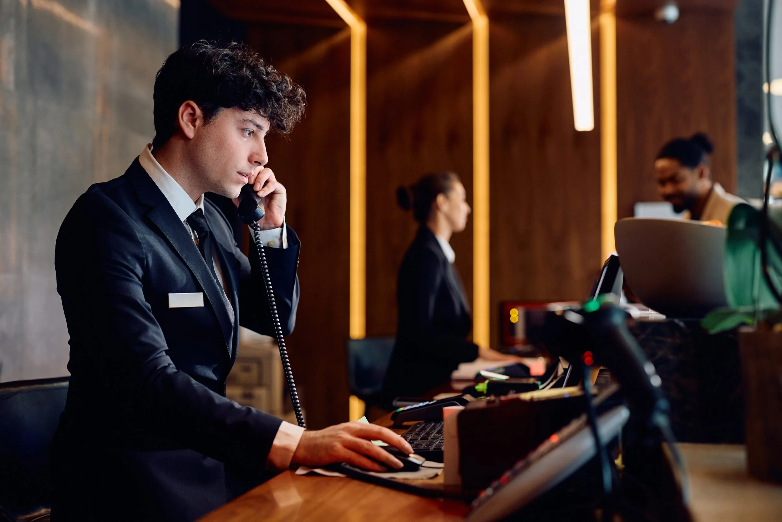 Young man in formal business attire using a telephone and working on a computer at a hotel reception or front desk. There are two women in uniforms working behind the counter.