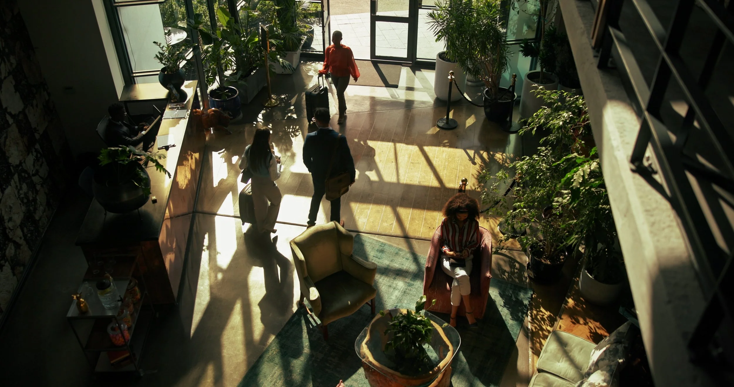Interior of a building lobby with people walking and sitting, sunlight streaming through large glass doors, plants, and furniture.