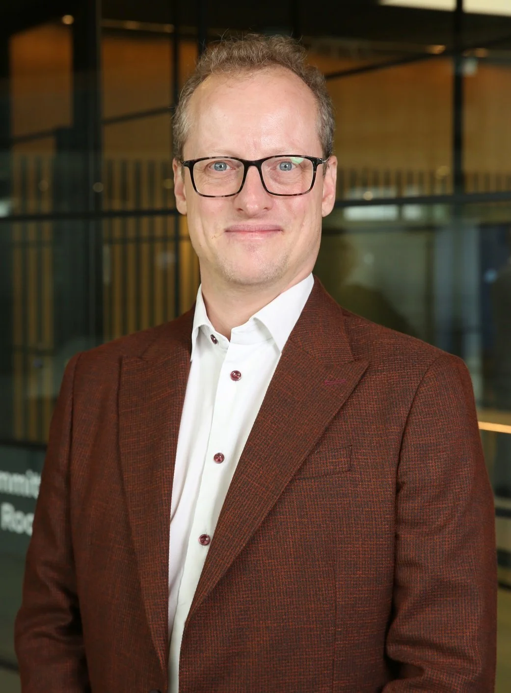 A portrait of Tjis Broeke wearing glasses, a white shirt, and a brown blazer, standing indoors with a modern office background.