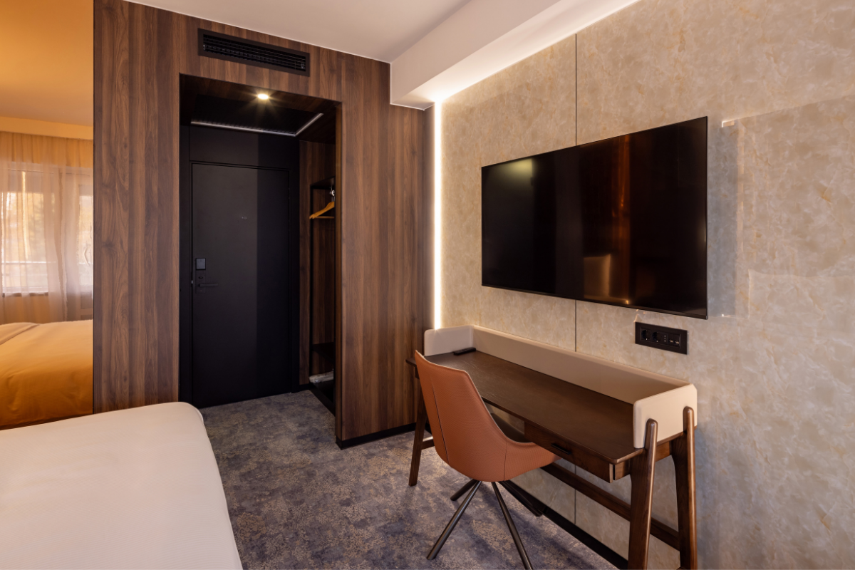 Hotel room with a wall-mounted TV, a small desk with a pink chair, a bed partially visible, and a wooden closet with an open door.