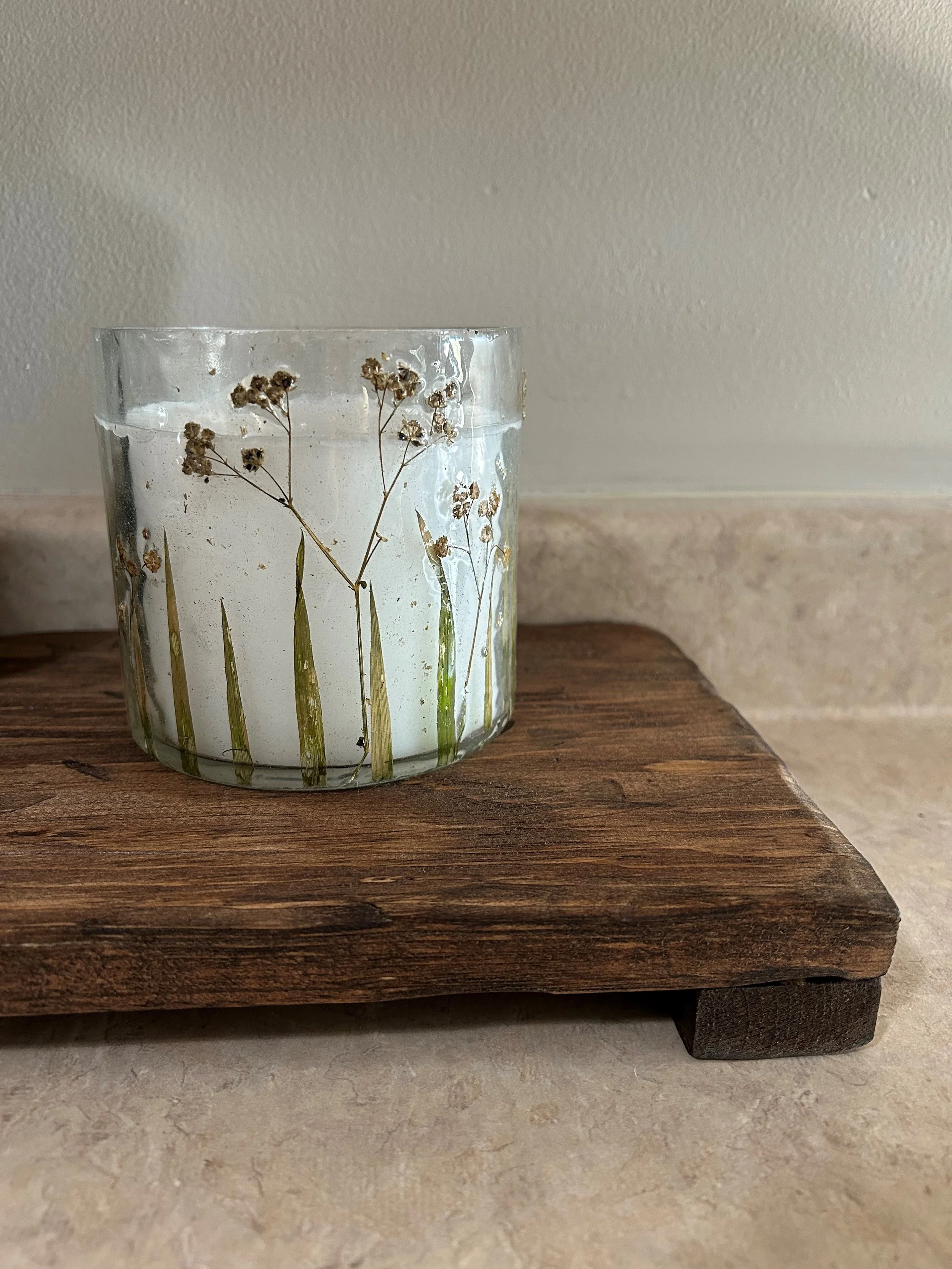 A glass candle holder with dried flowers inside, sitting on a wooden surface.