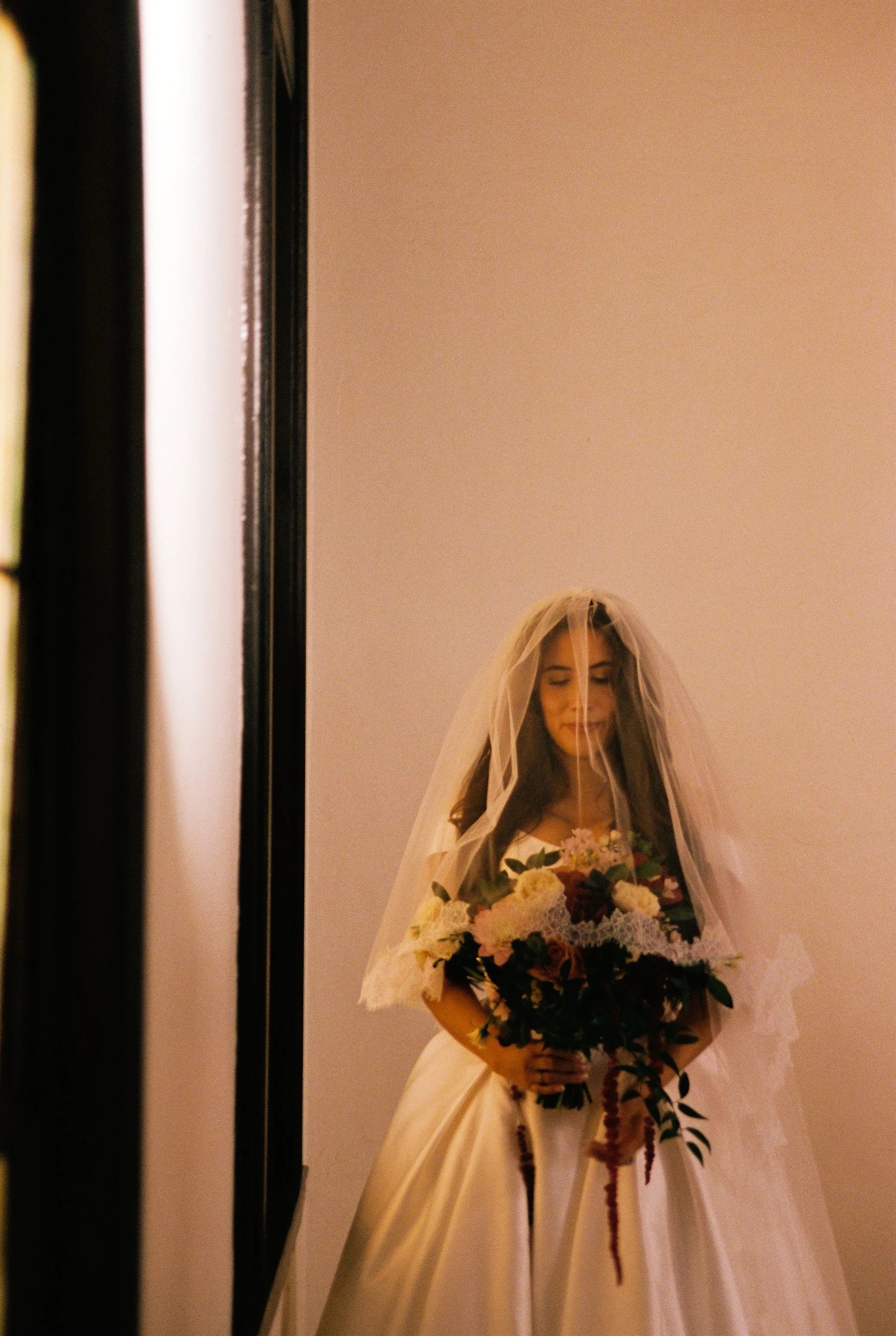 A woman in a wedding dress with a veil, holding a bouquet of flowers, reflected in a mirror.