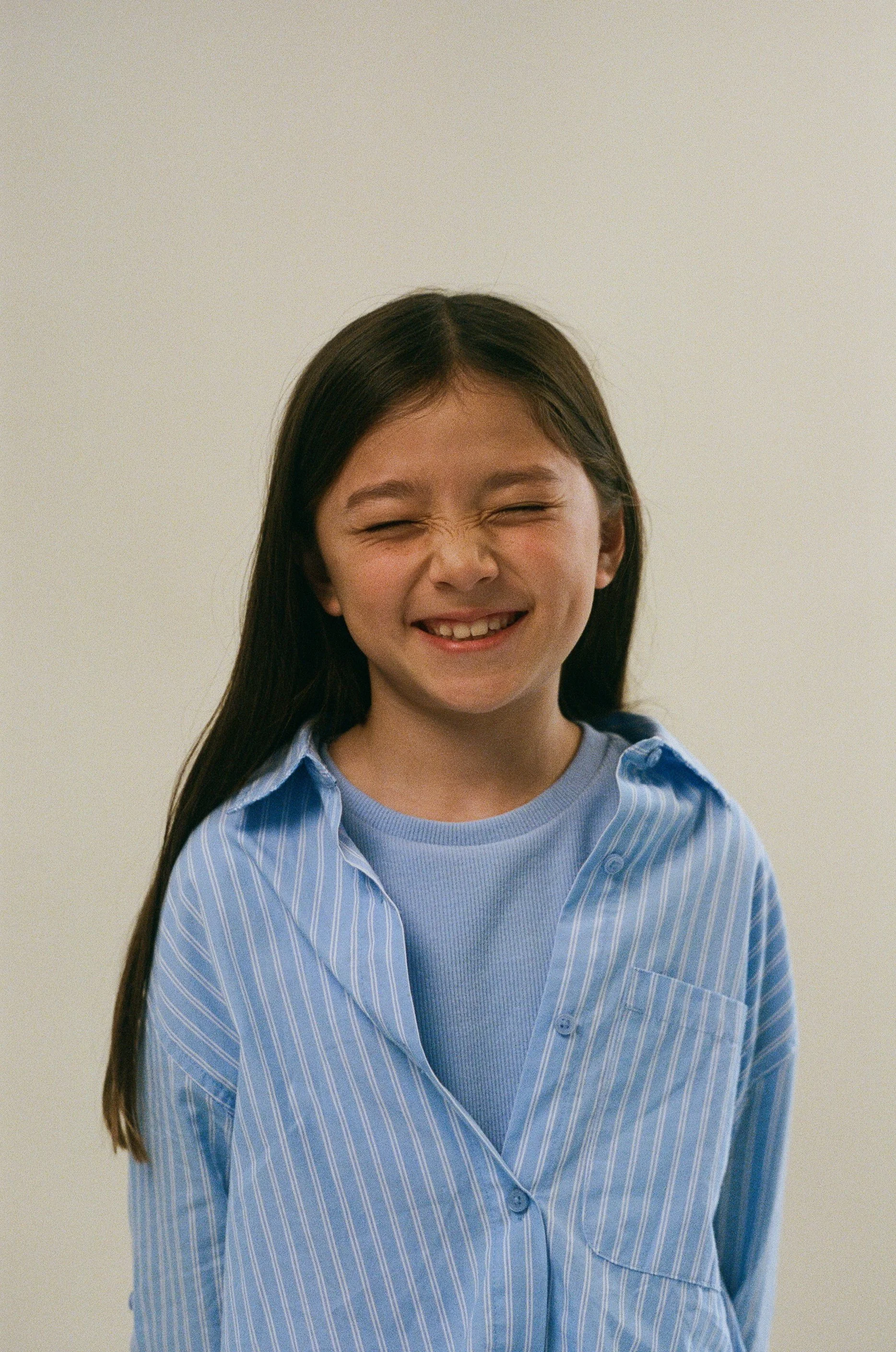 Smiling girl with long dark hair wearing a blue striped button-up shirt over a gray t-shirt, standing against a plain beige background.