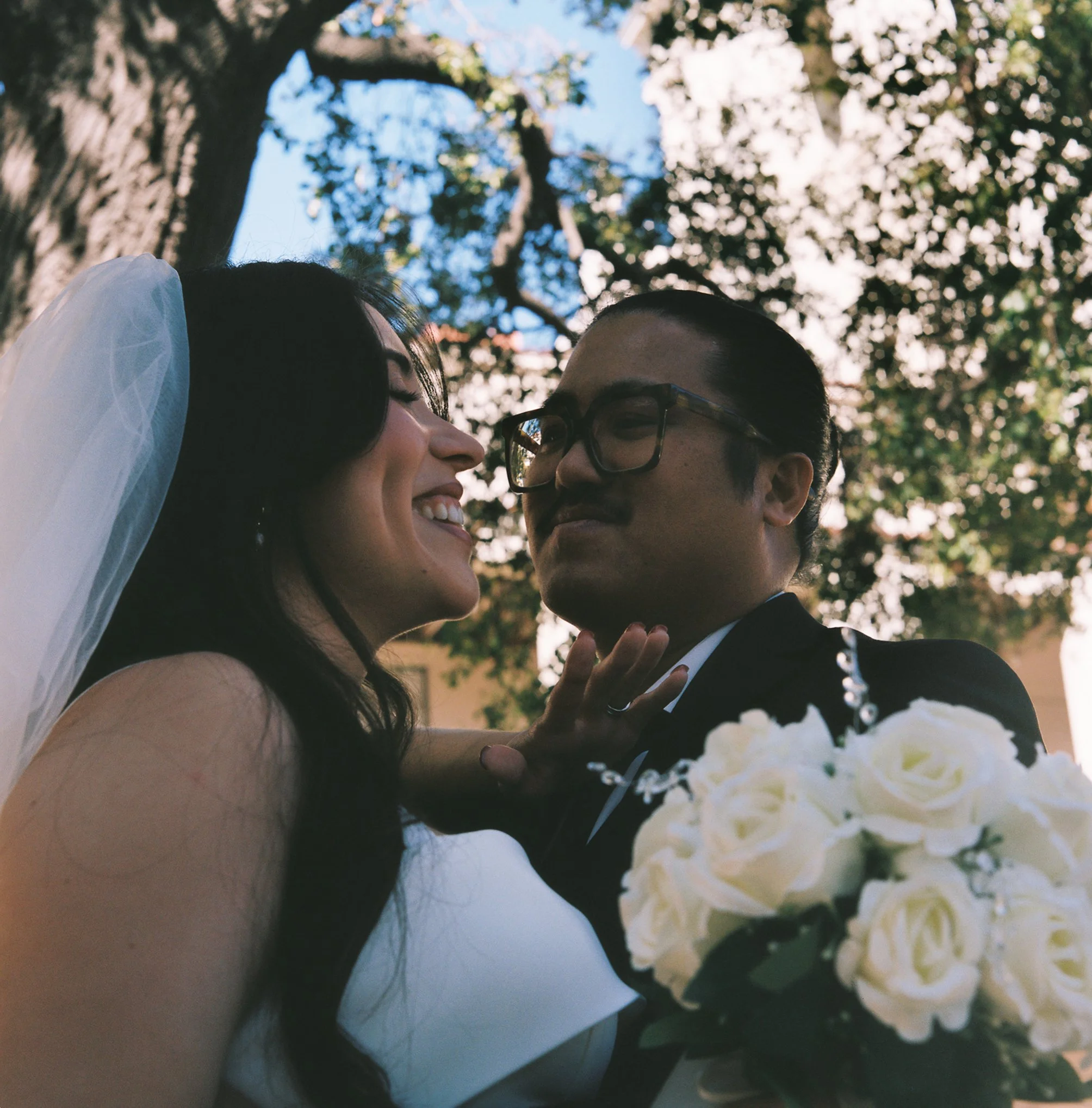 Close-up of a woman and a man smiling and embracing each other outdoors on a sunny day, with large trees and blue sky in the background.
