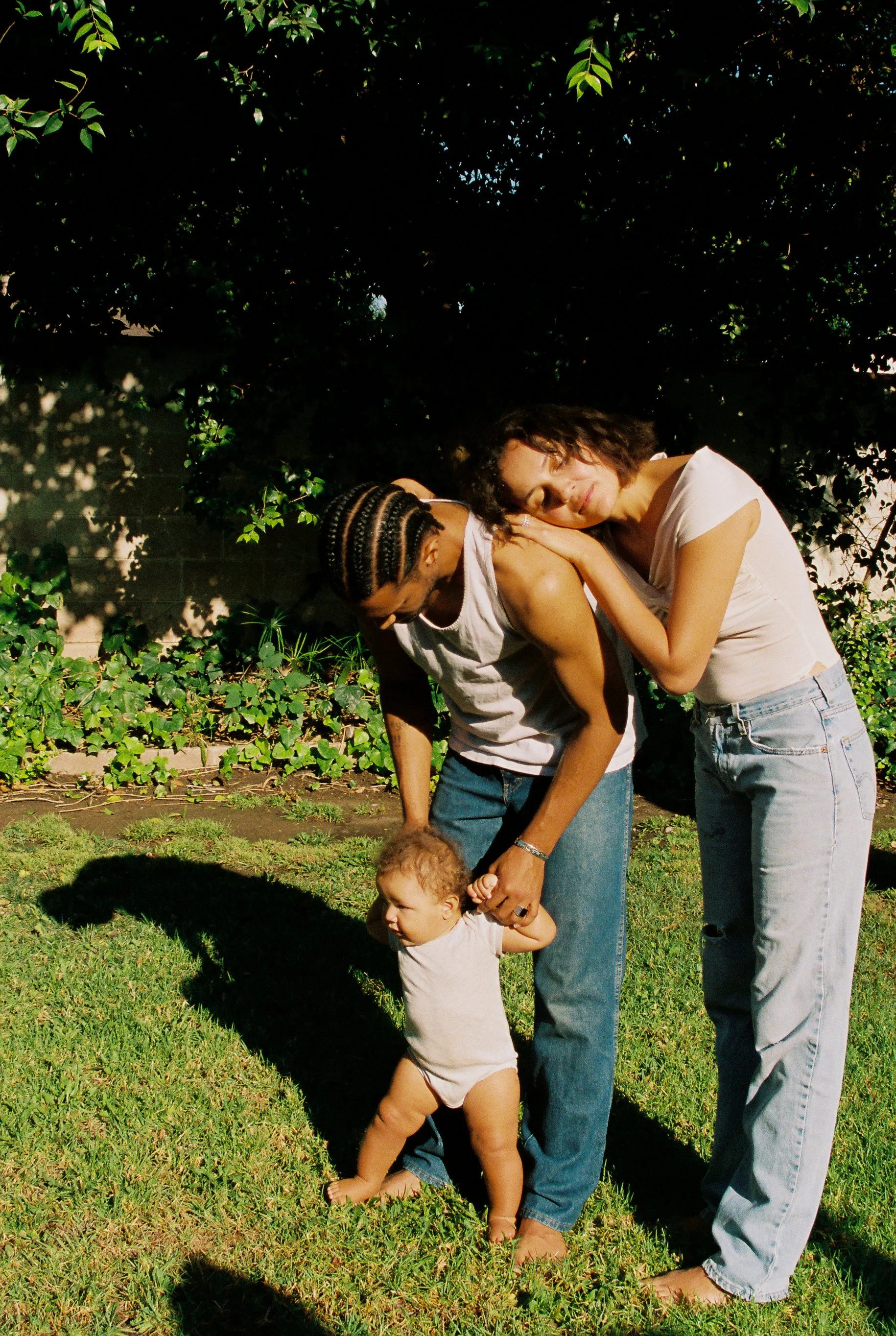Two women and a young child outdoors on grass with a fence and leafy trees in the background. One woman has curly hair and is resting her head on the other woman’s shoulder. The woman with the curly hair is wearing a light-colored t-shirt and jeans. 