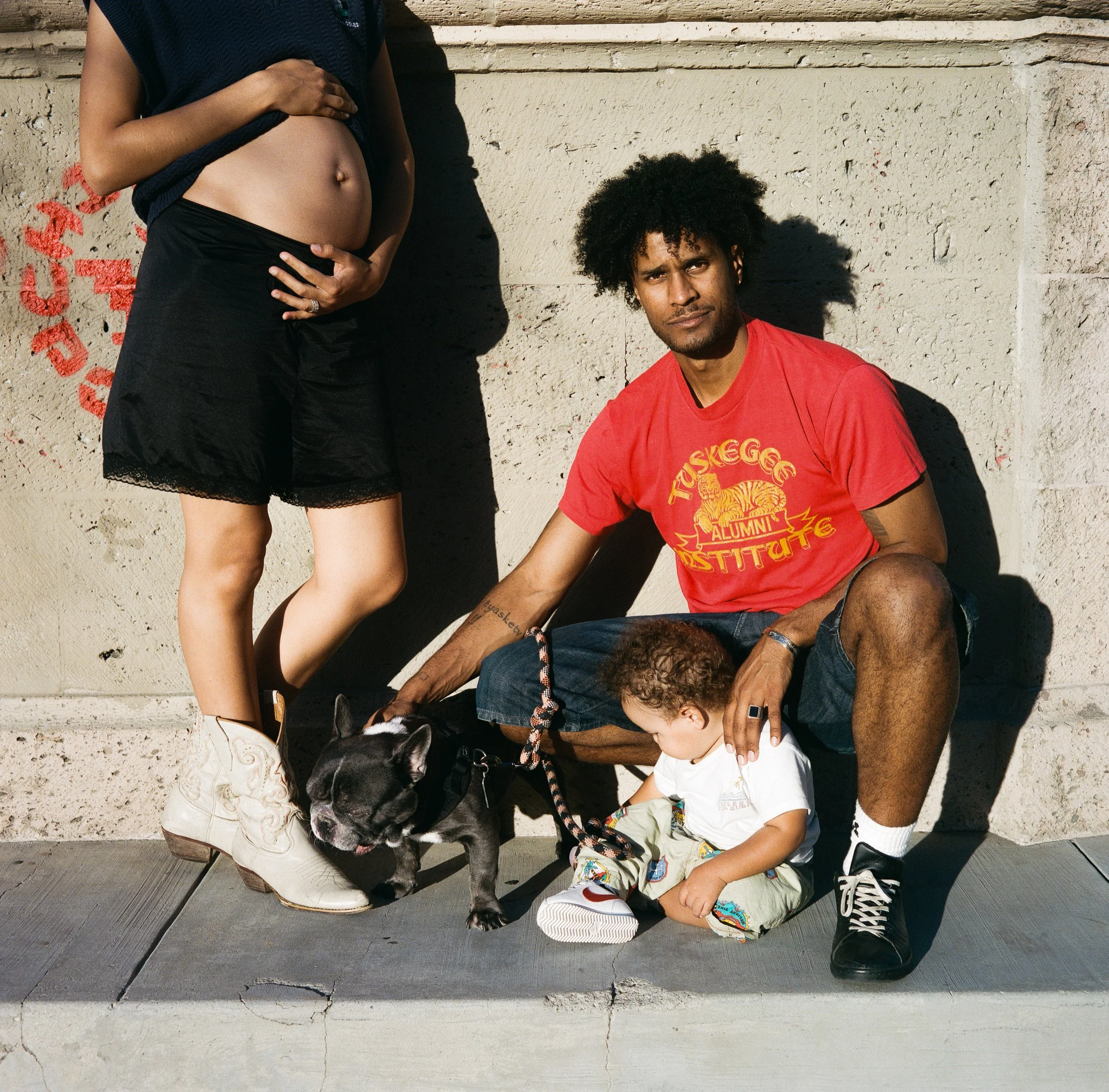 A black and white French Bulldog puppy with a red harness standing next to a seated young boy wearing colorful shorts, white sneakers, and a white T-shirt. The puppy is looking to the right while the boy is looking down with his eyes closed. There are two adults: one standing with a hand on a pregnant woman's belly and wearing a black skirt and a dark top, and the other sitting with a man with curly hair wearing a red T-shirt with yellow text and graphics. They are outdoors against a beige wall with red graffiti and textured surface.
