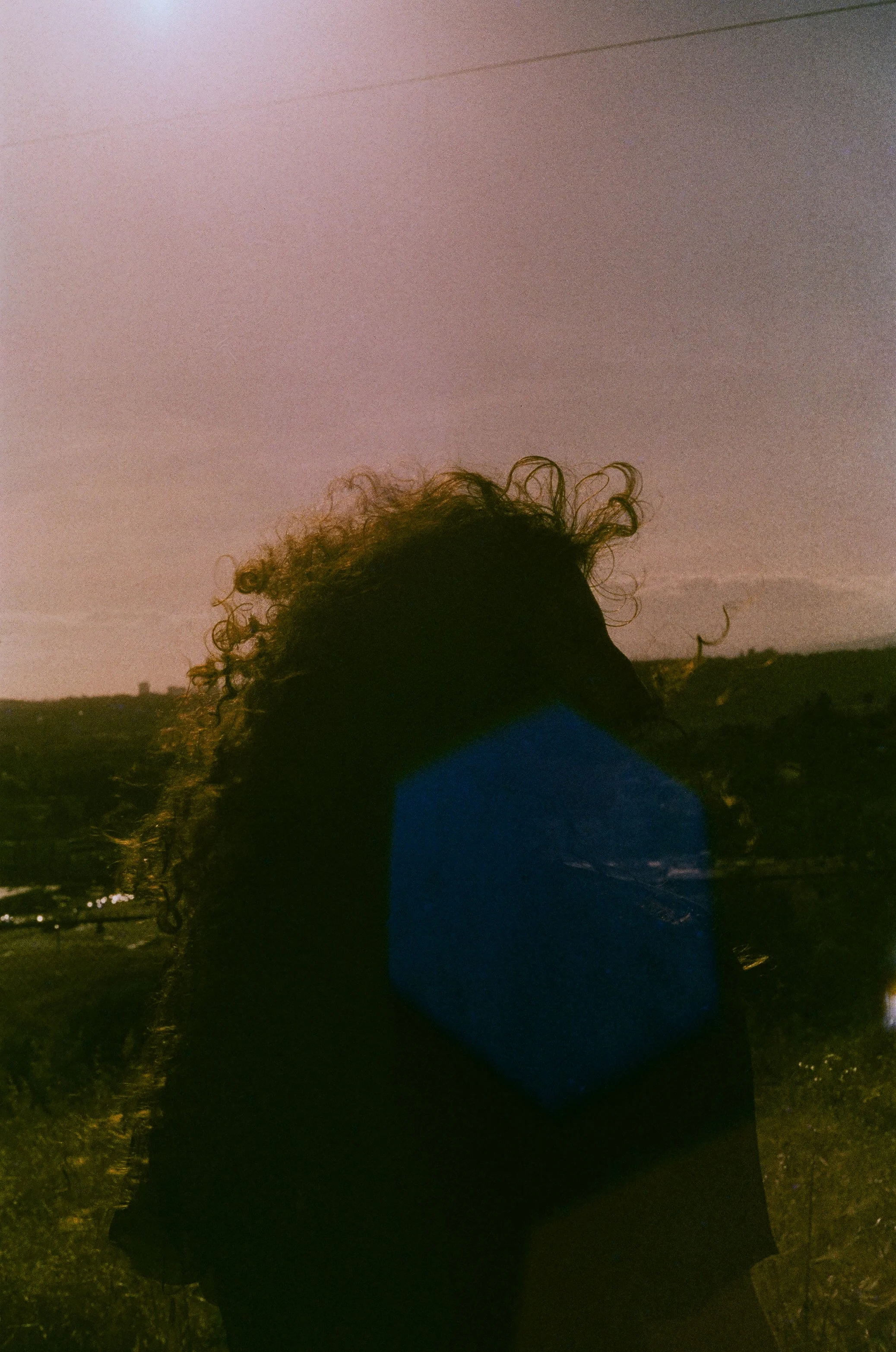 A person with curly hair standing outdoors during sunset or sunrise, with a landscape and power lines in the background.