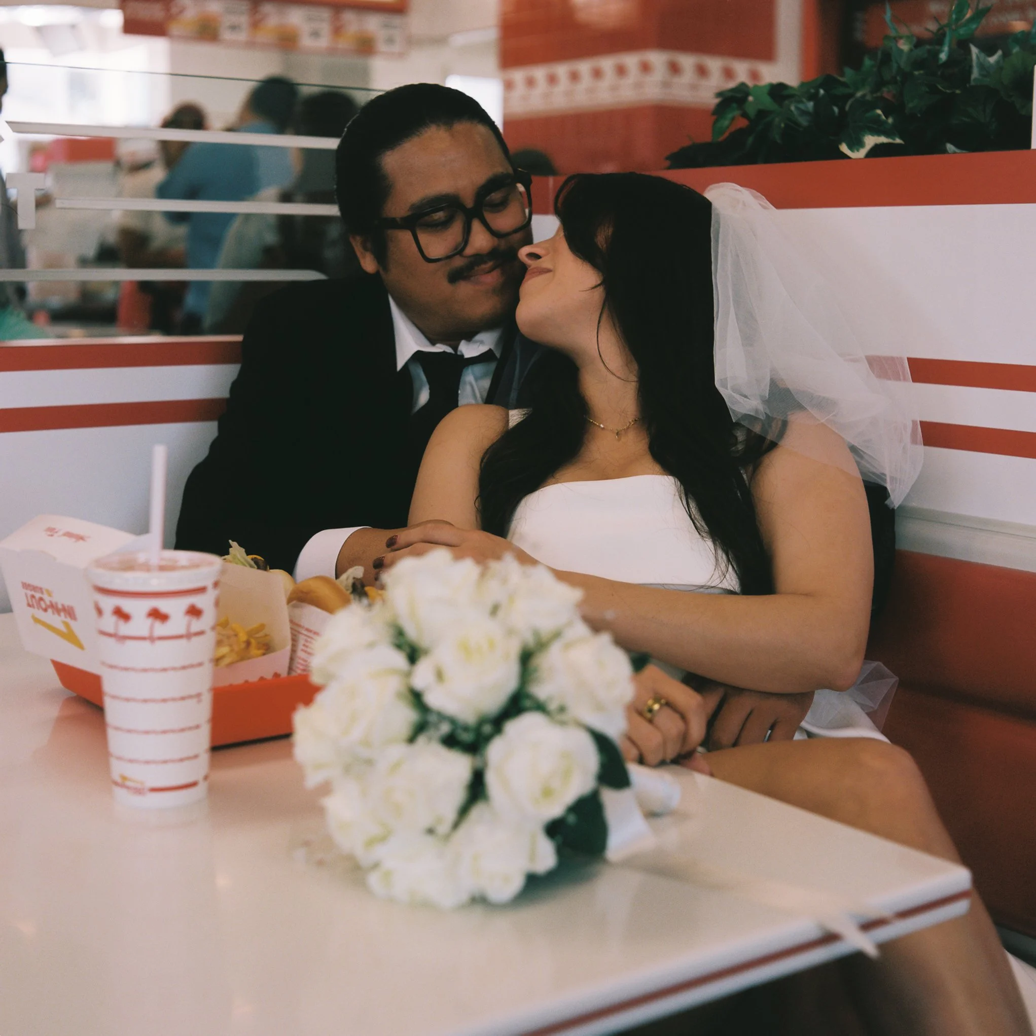 A man and woman share a kiss at a fast food restaurant table, with trays of food including fries and soda in the foreground and other customers in the background.