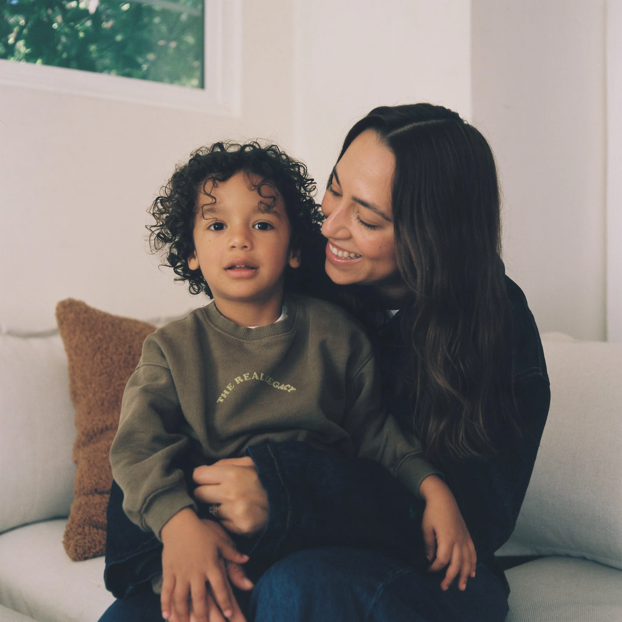 A woman smiling and holding a young child with curly hair, sitting indoors near a window.