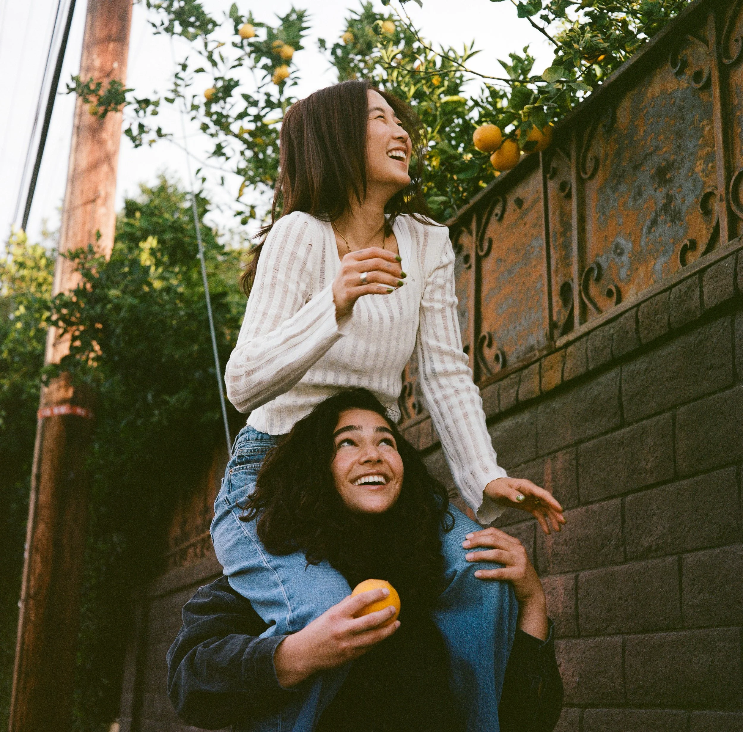 Two women, one sitting and one standing on her shoulders, both smiling and holding lemons, in an outdoor setting with trees and a rusty wall.