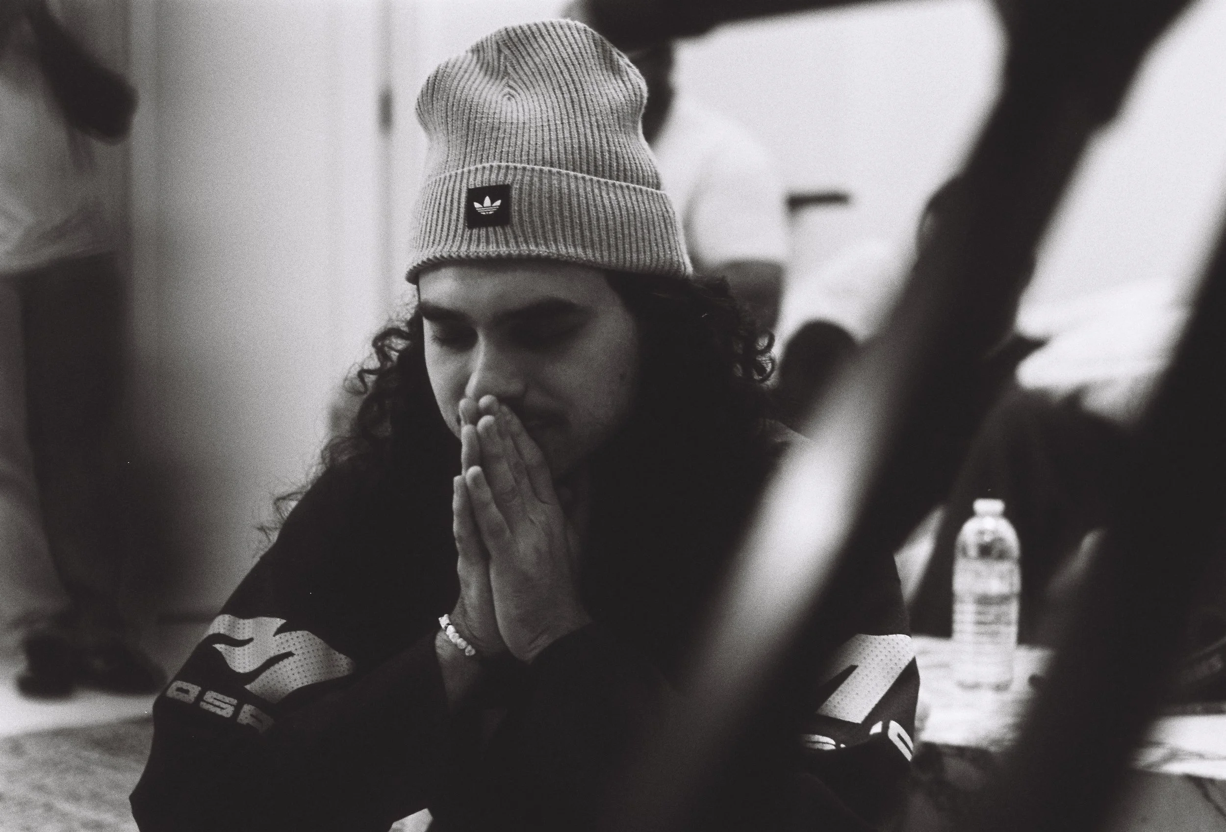 A woman wearing a knit beanie with an Adidas logo, praying with her hands pressed together during a gathering.