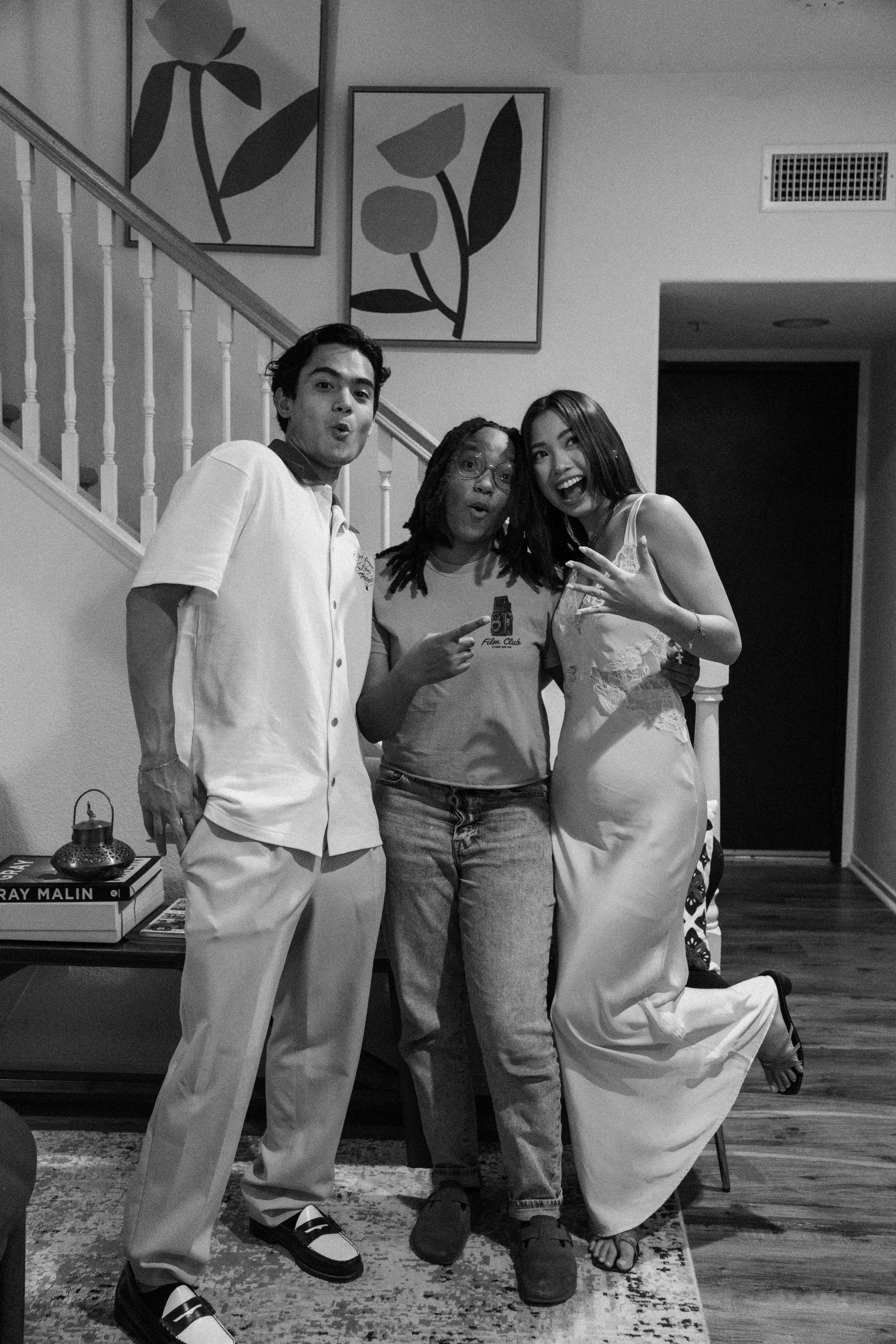 Three friends having fun indoors. A man in a white shirt, a woman in a white dress, and another woman in a t-shirt and jeans. They are smiling, making expressive faces, and posing together. The background shows artwork on the wall, books, and a rug on the floor.