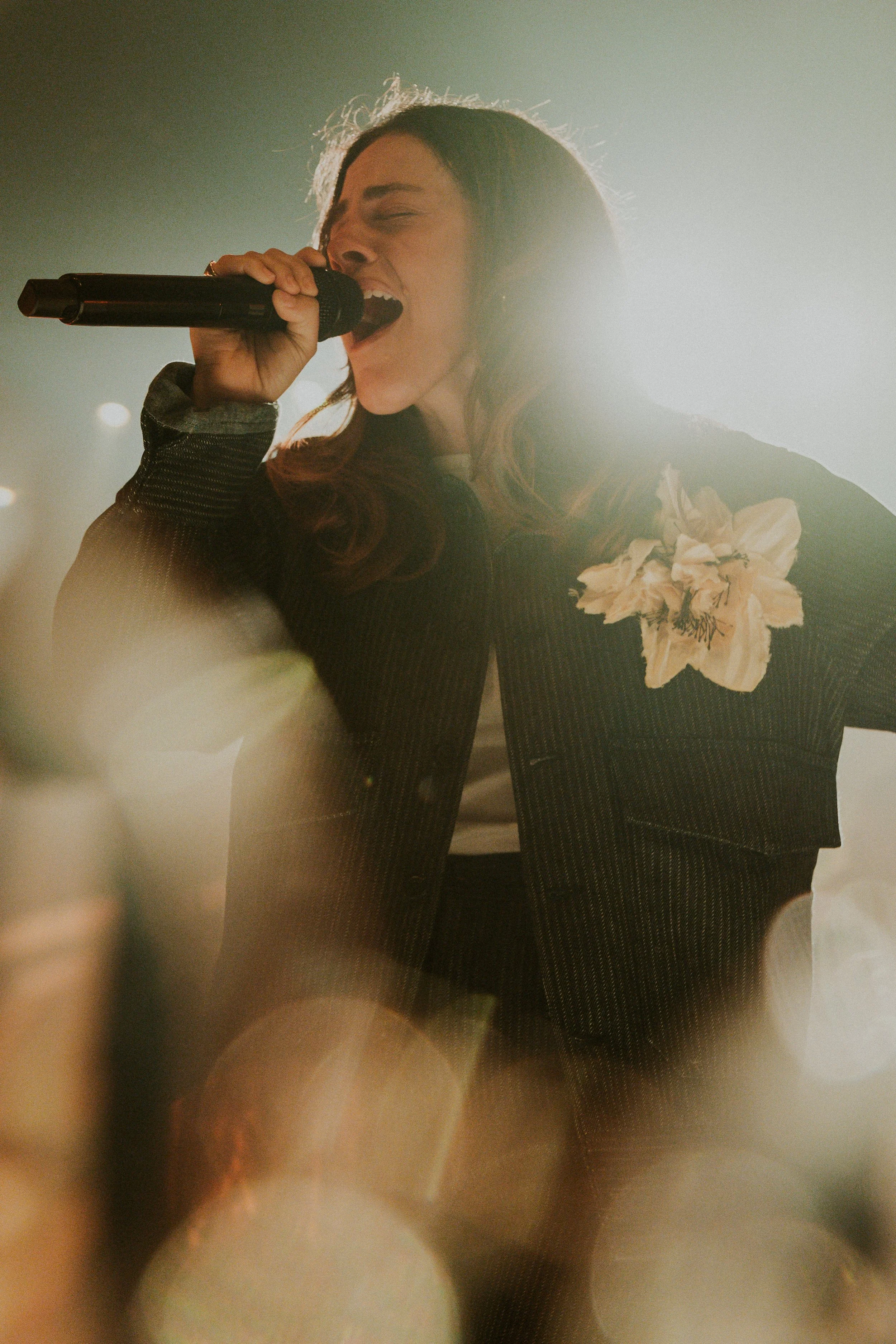 A woman singing passionately into a microphone during a performance, wearing a black blazer with a floral decoration on it.