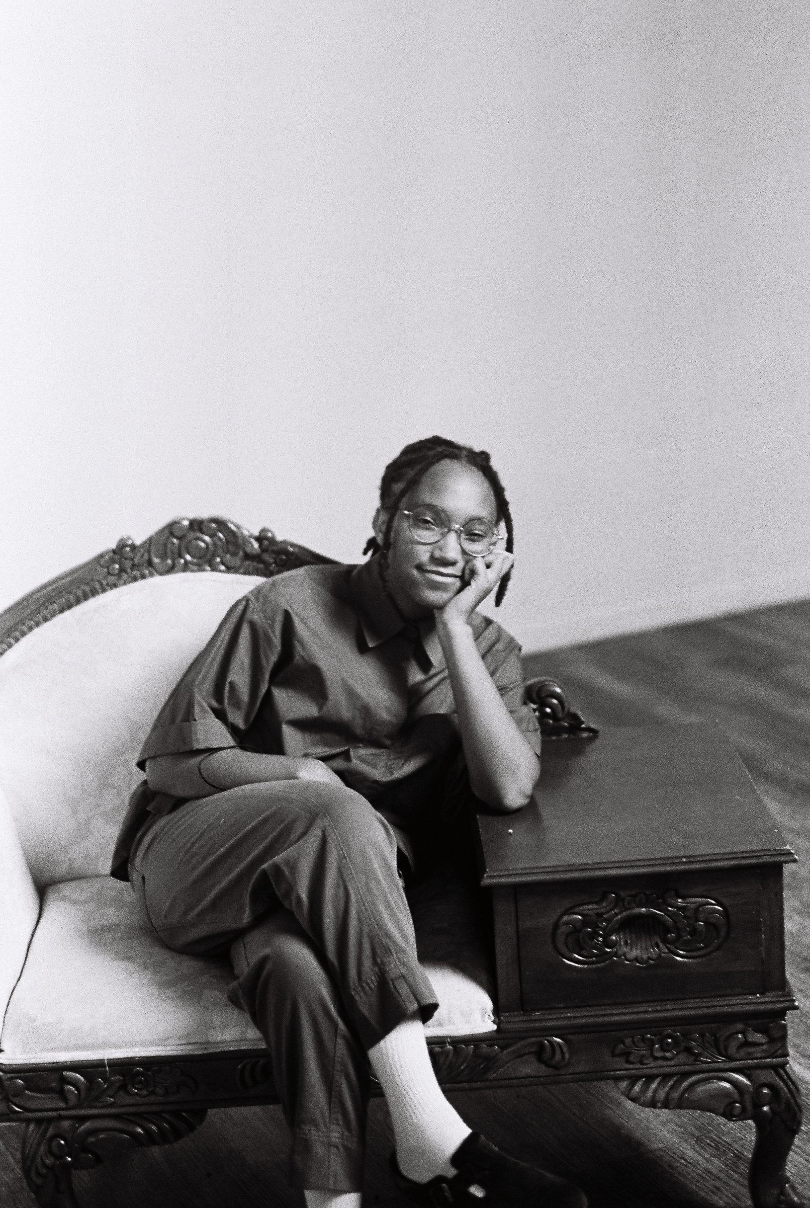 A young person with glasses and dreadlocks sitting on an ornate wooden bench, resting their head on their hand, and smiling in a room with plain walls.