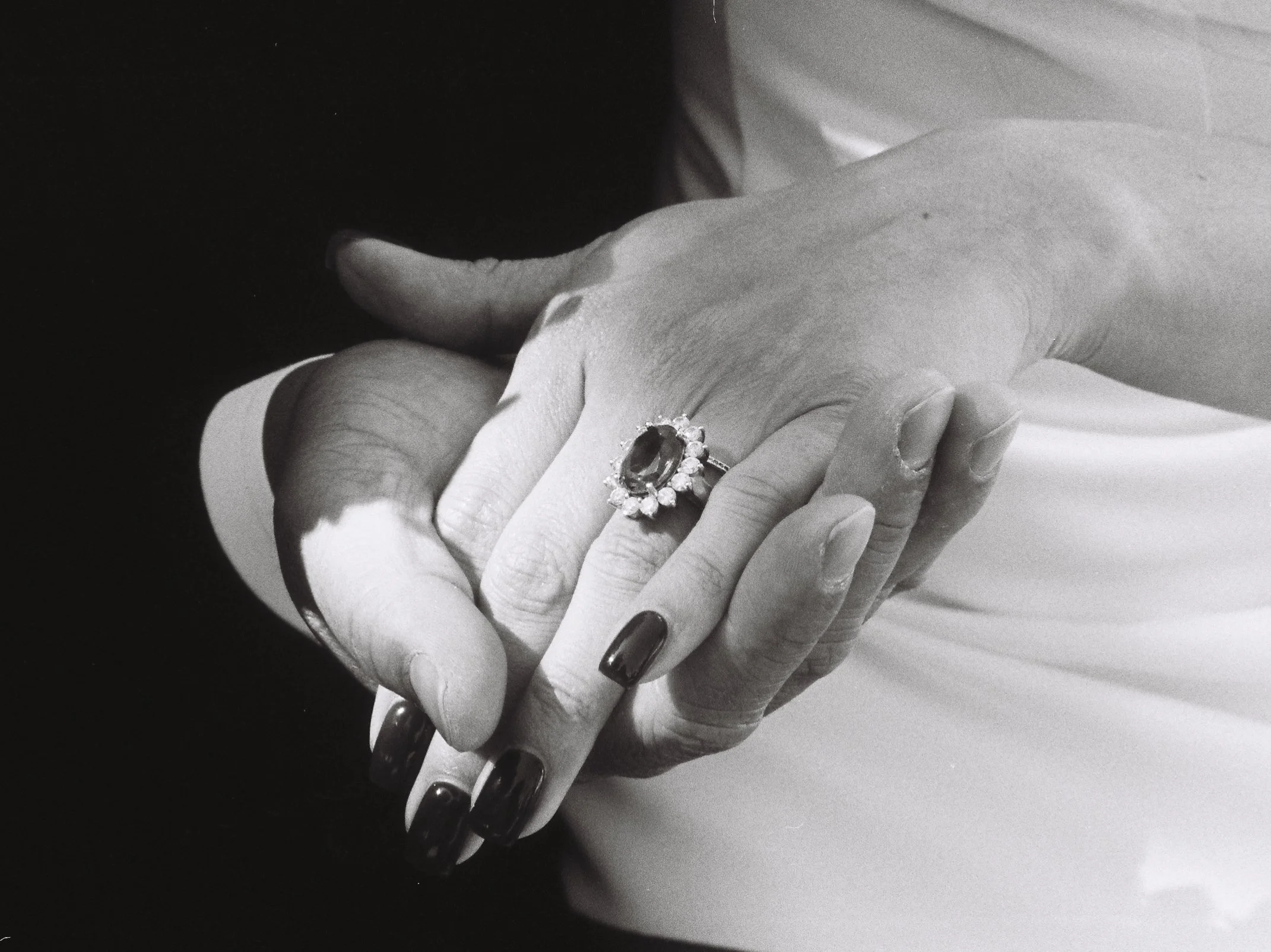 Black and white photograph of two hands intertwined, one wearing a large ring with a central dark gemstone surrounded by smaller stones, and polished black nails.
