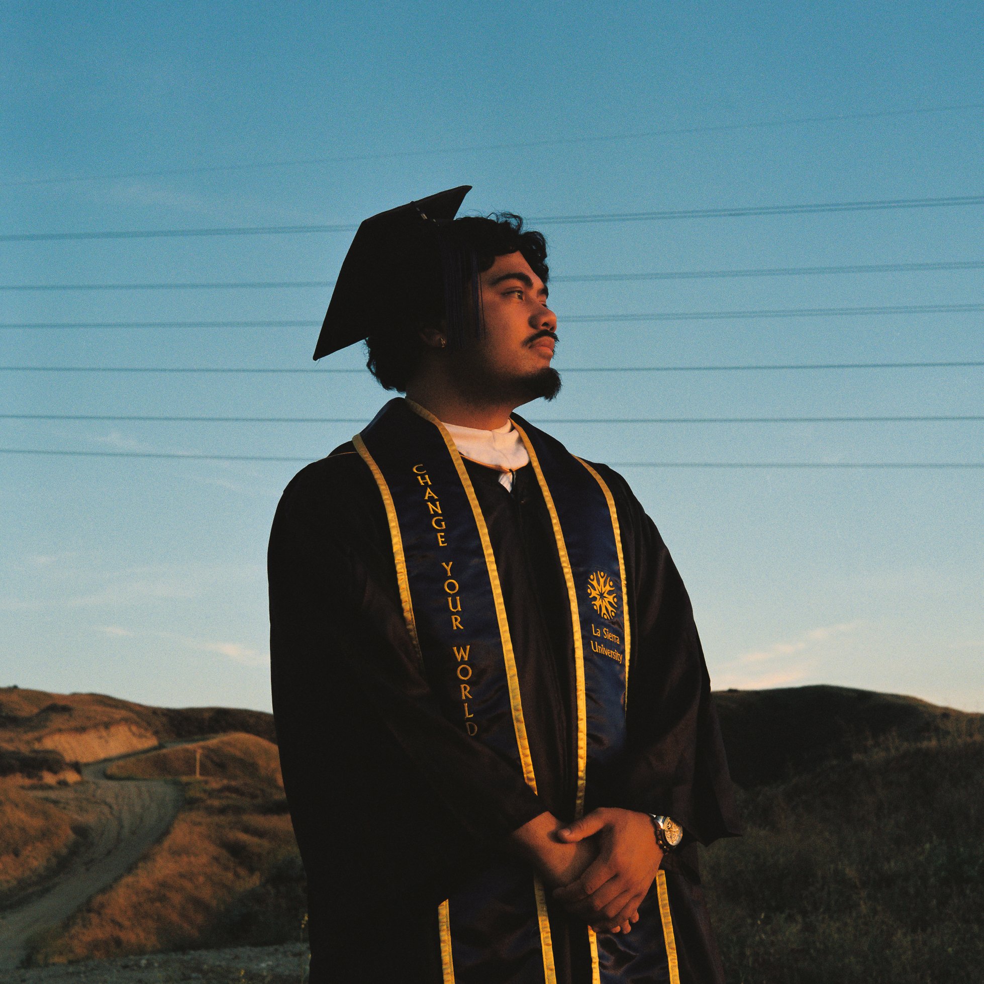 A young man in a black graduation gown with yellow trim wearing a graduation stole and cap stands outdoors during sunset or dusk, with power lines and hills in the background.