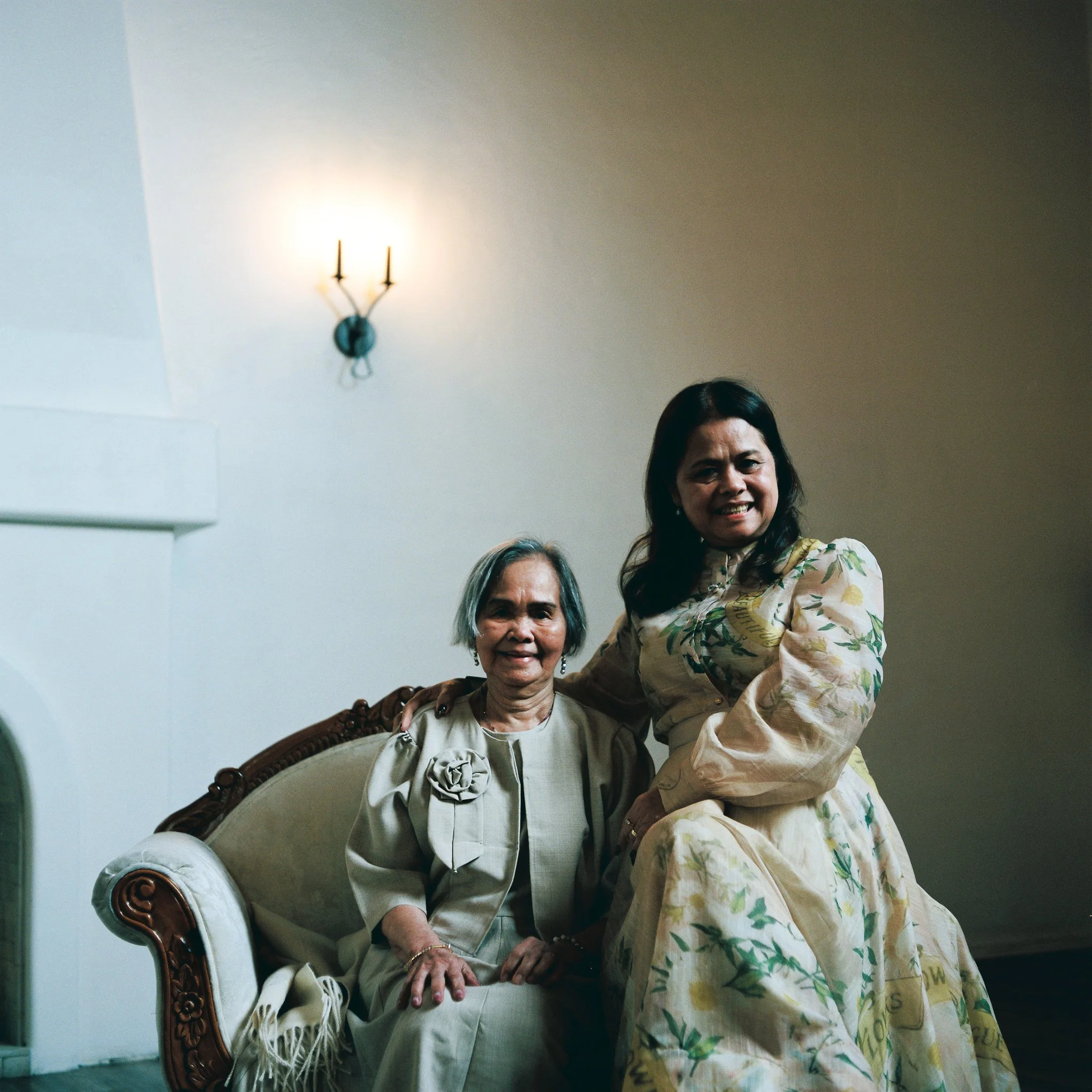Two women smiling, one seated in an ornate armchair and another standing beside her with one arm around her shoulder, in a room with plain walls and a wall-mounted chandelier.