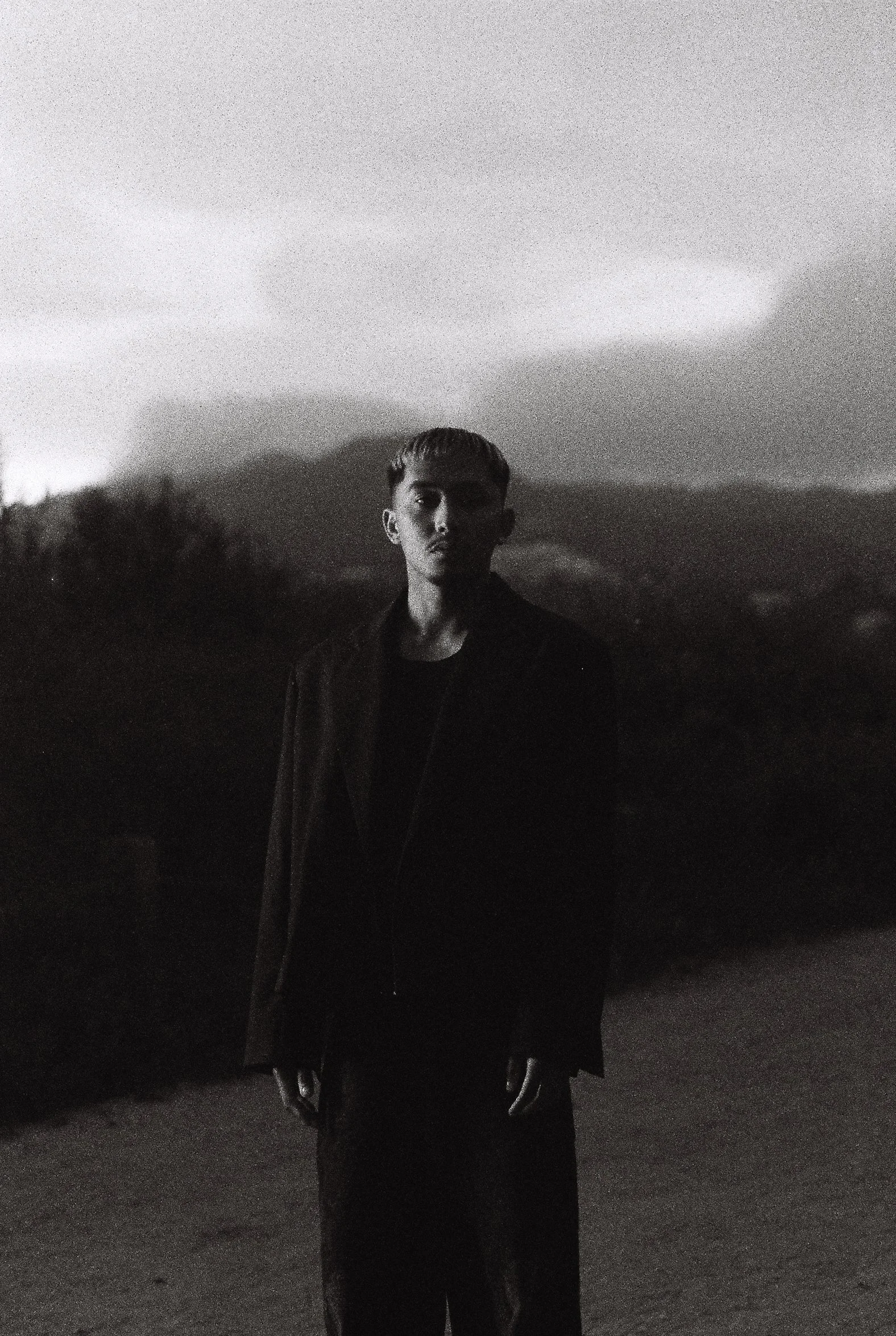 A black and white photo of a young man with short hair, standing outdoors at dusk or night, with mountains or hills in the background.
