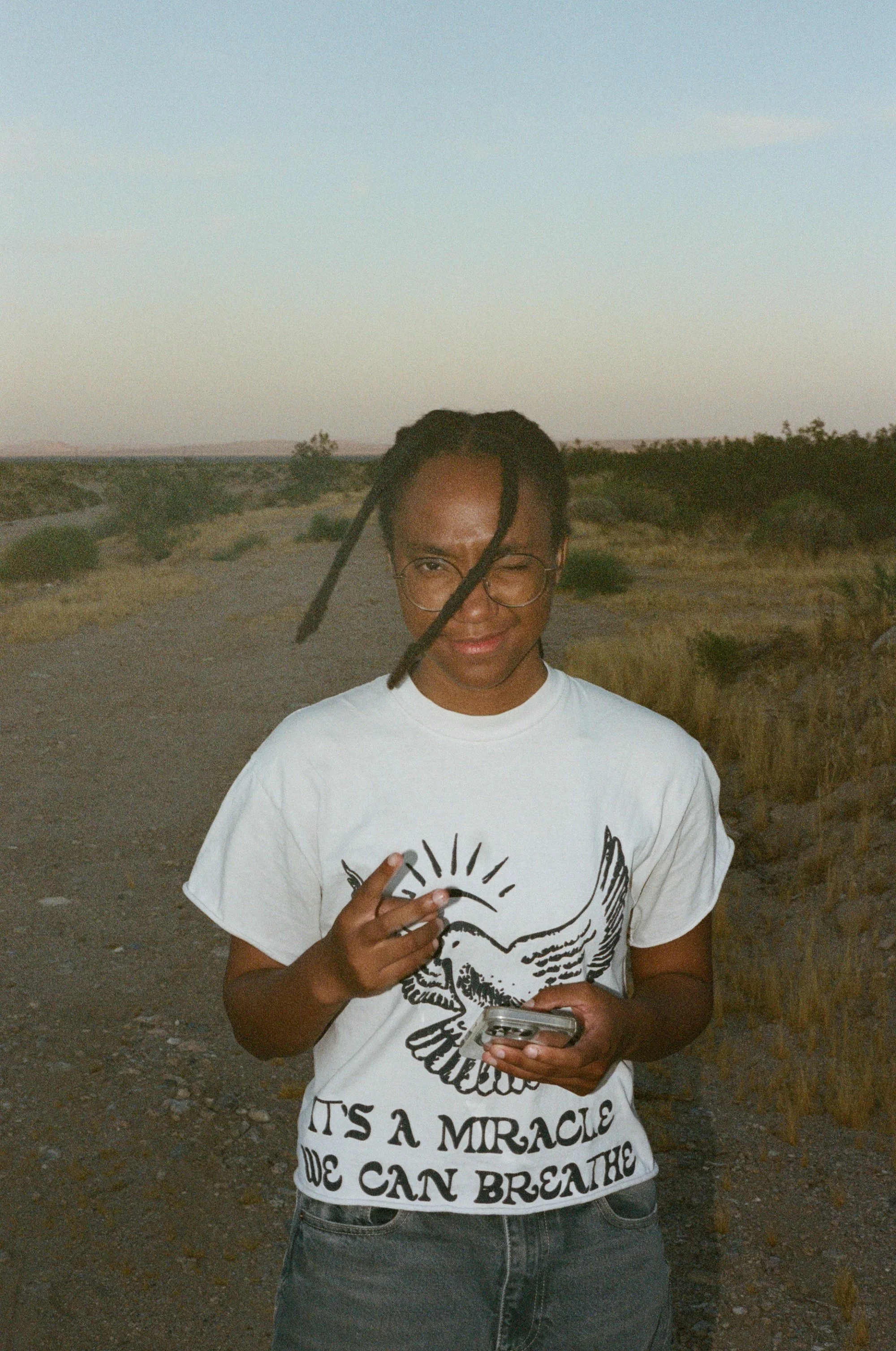 Young person with glasses and dreadlocks winking, holding a phone and a cigarette, standing outdoors in a desert landscape with dry bushes, wearing a white graphic T-shirt that says 'It's a miracle we can breathe'.