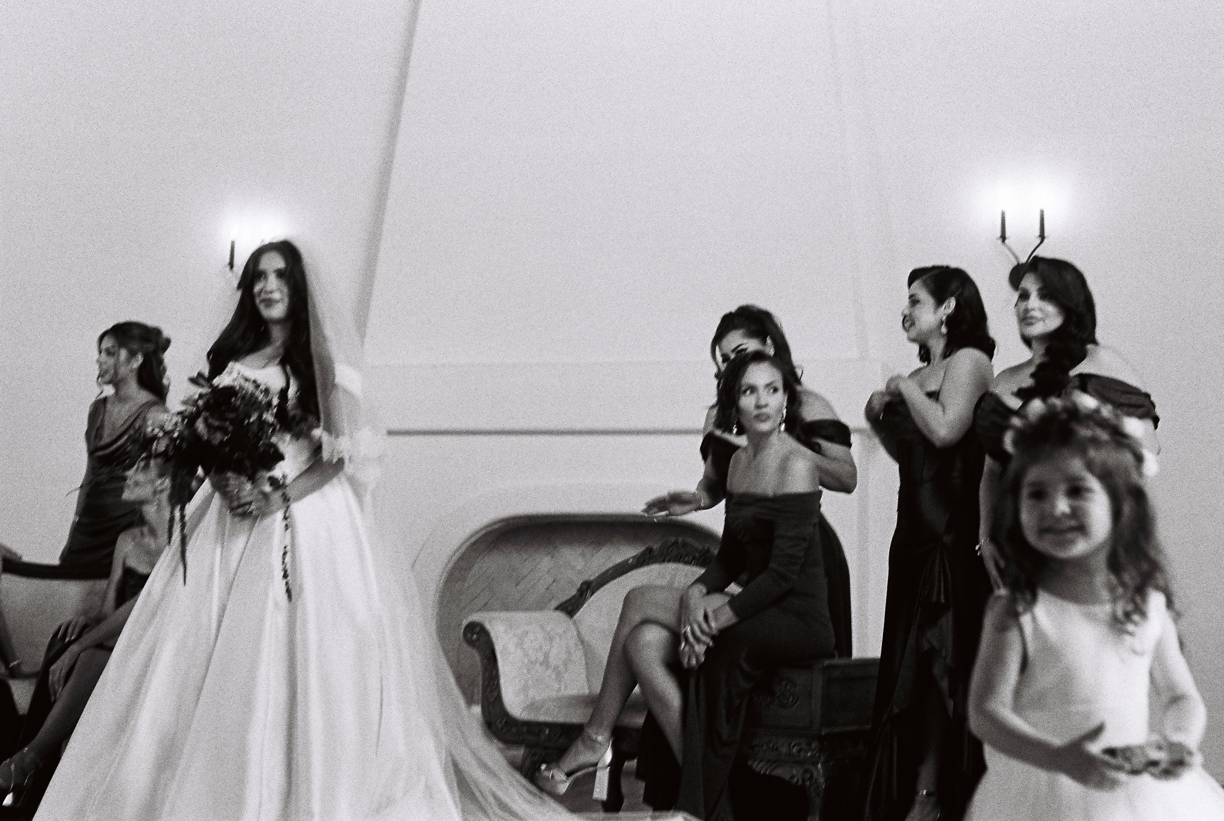 Group of women in formal dresses gathered at a social event, some smiling and engaging with each other, in an indoor setting with elegant interior decor.