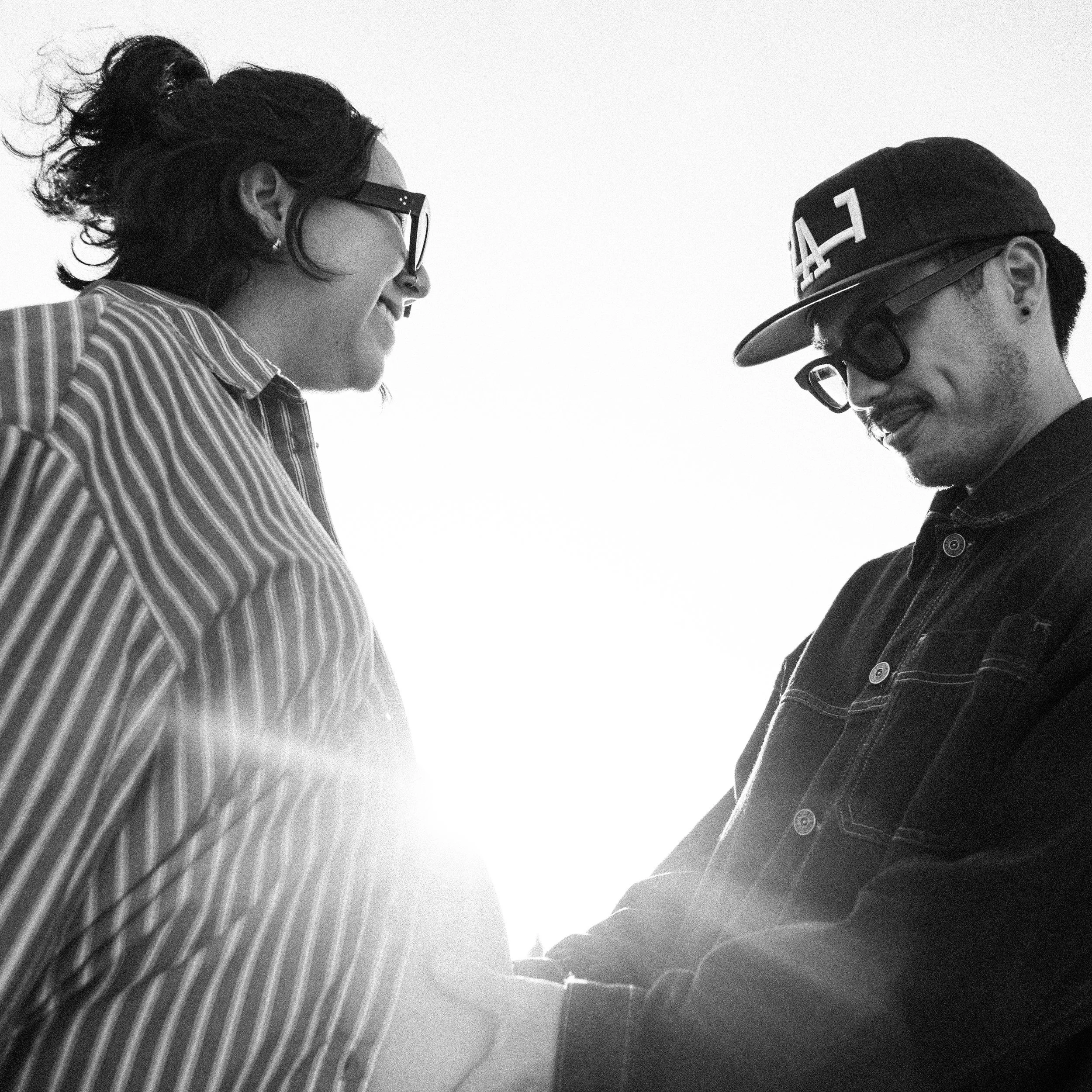 Two people smiling and looking at each other outdoors, wearing sunglasses and casual clothing, with a clear sky in the background.