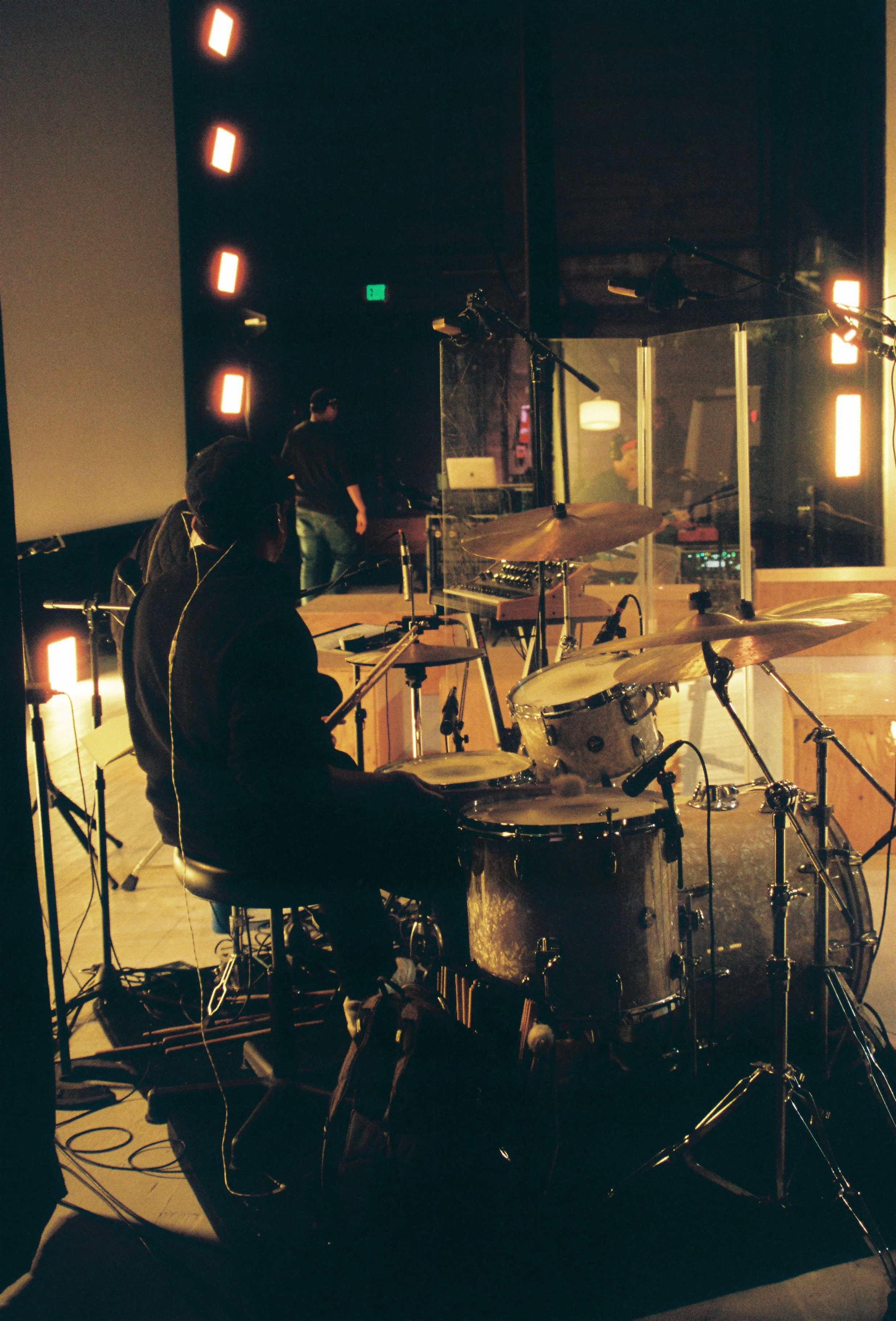 A recording studio with musicians and sound equipment, including drums, microphones, and audio gear, with a dark background and warm lighting.