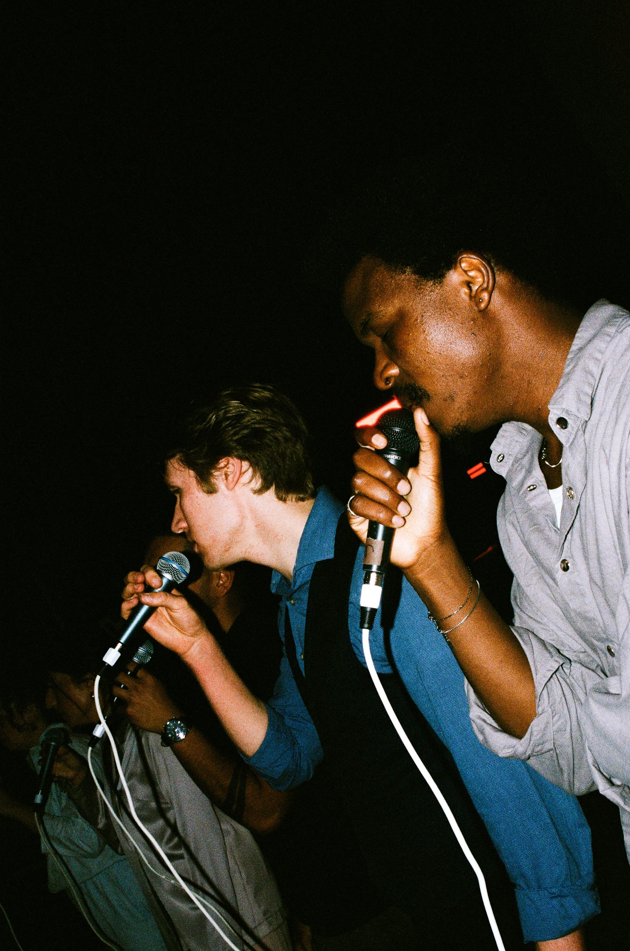 Multiple people are holding microphones and singing or speaking into them at an event in a dark setting.