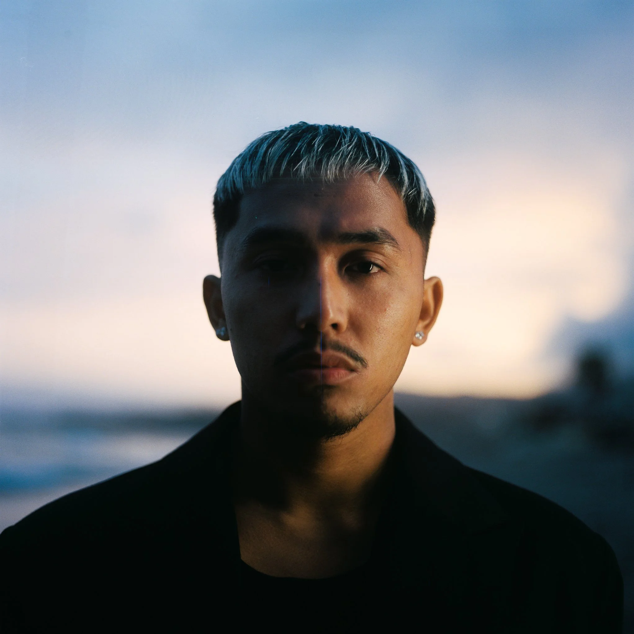 Close-up of a young person with short, two-tone hair and earrings, looking towards the camera with a sunset or twilight background.