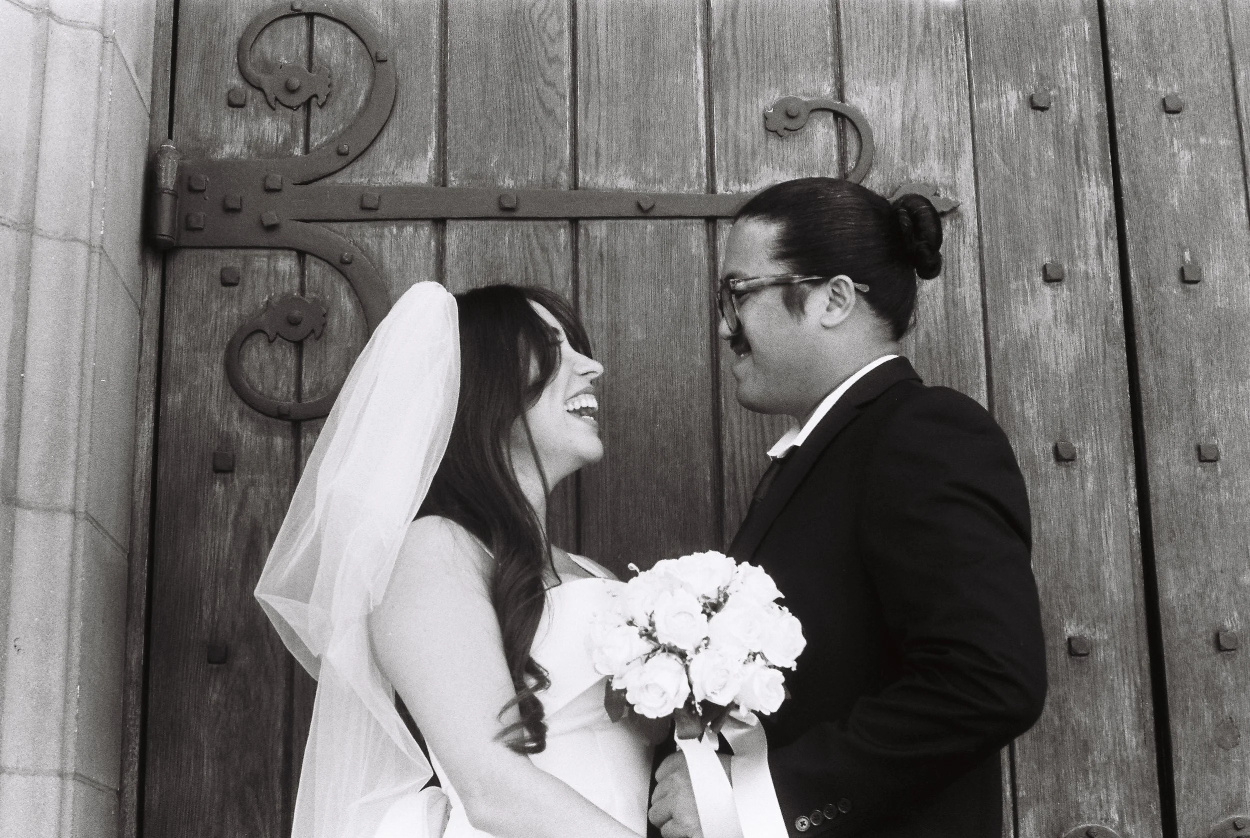 A black and white photo of a bride and groom standing close together in front of a large wooden door with decorative metal hinges. The bride is wearing a wedding dress, veil, and holding a bouquet of flowers. The groom is dressed in a dark suit with glasses, and they are both smiling at each other.