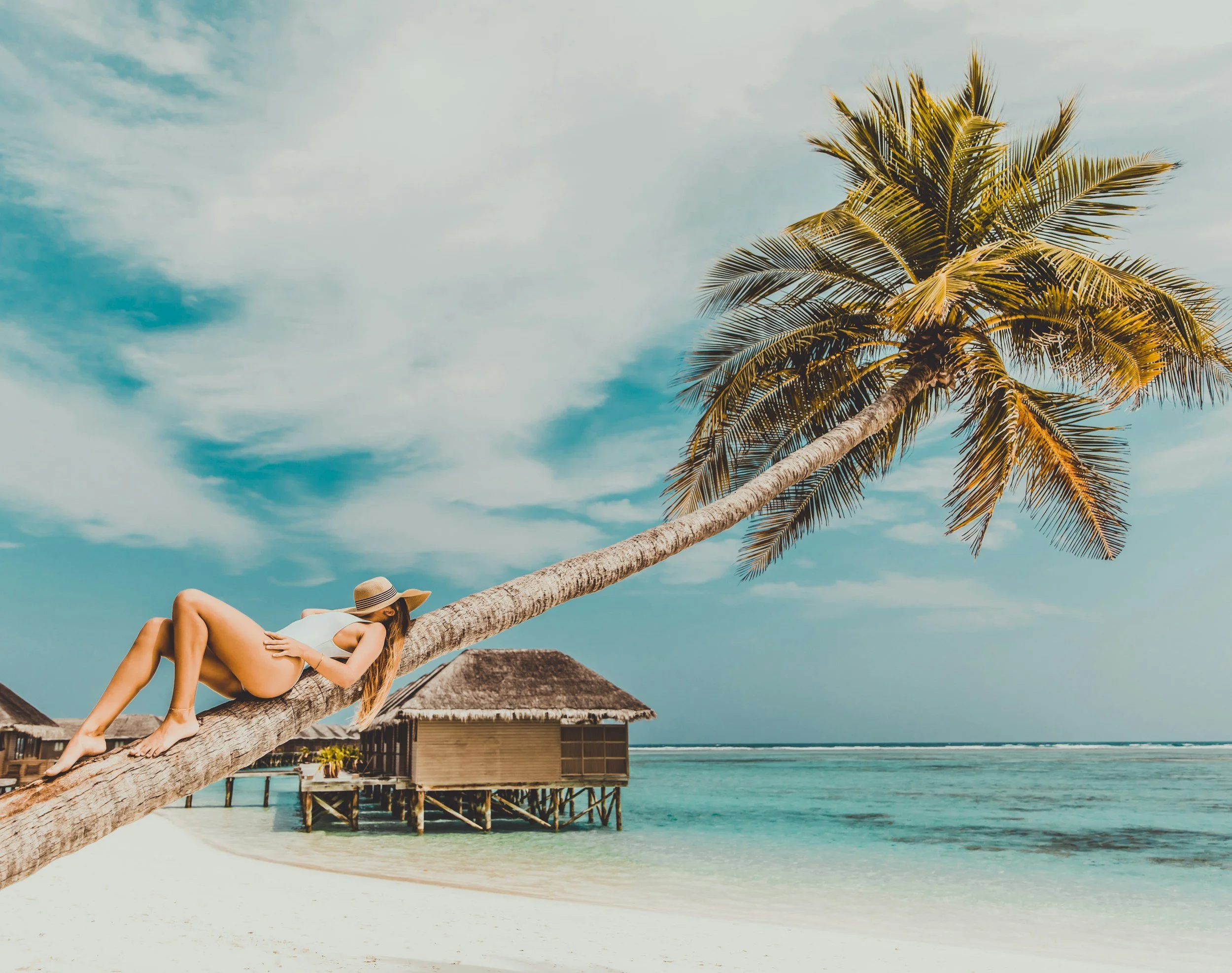 A woman lying on a leaning palm tree that extends over a beach with overwater bungalows in the background, under a partly cloudy sky at a tropical seaside location.