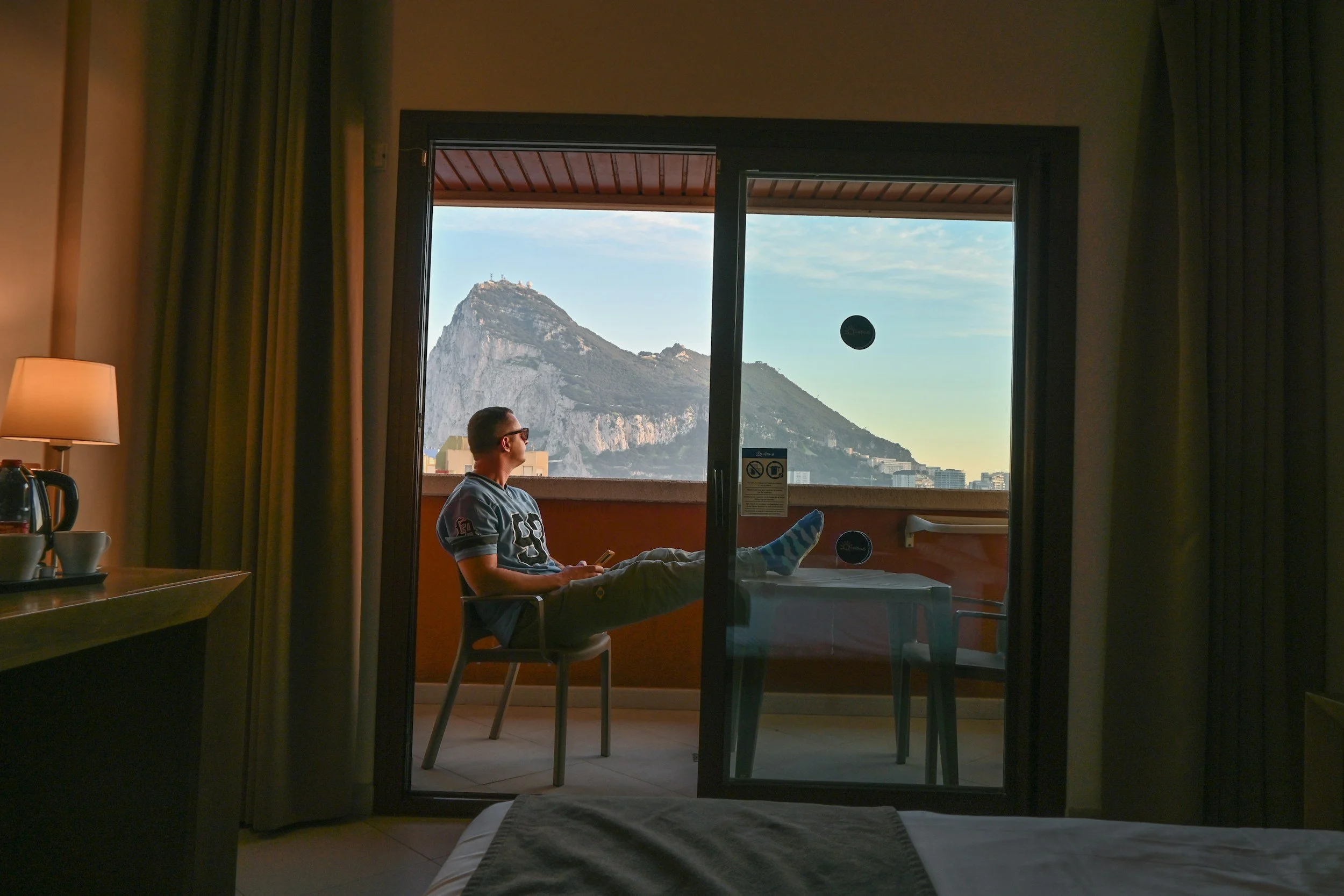 A person sitting on a hotel balcony, looking at the mountain and city view outside, with a mountain and buildings in the background.