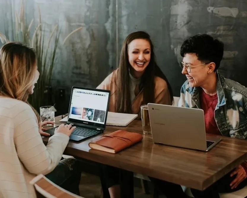 Three people sitting at a wooden table, smiling and working on laptops, with notebooks and drinks on the table in a casual, modern setting.