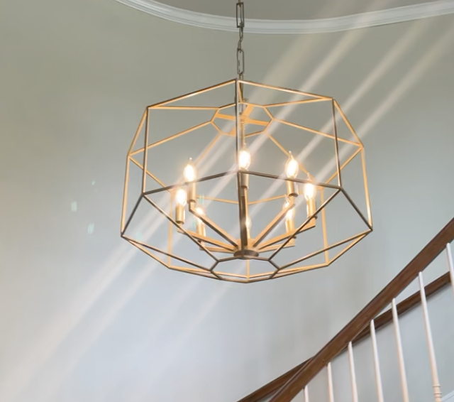 Modern geometric gold chandelier with four candles hanging from a white ceiling near a staircase.