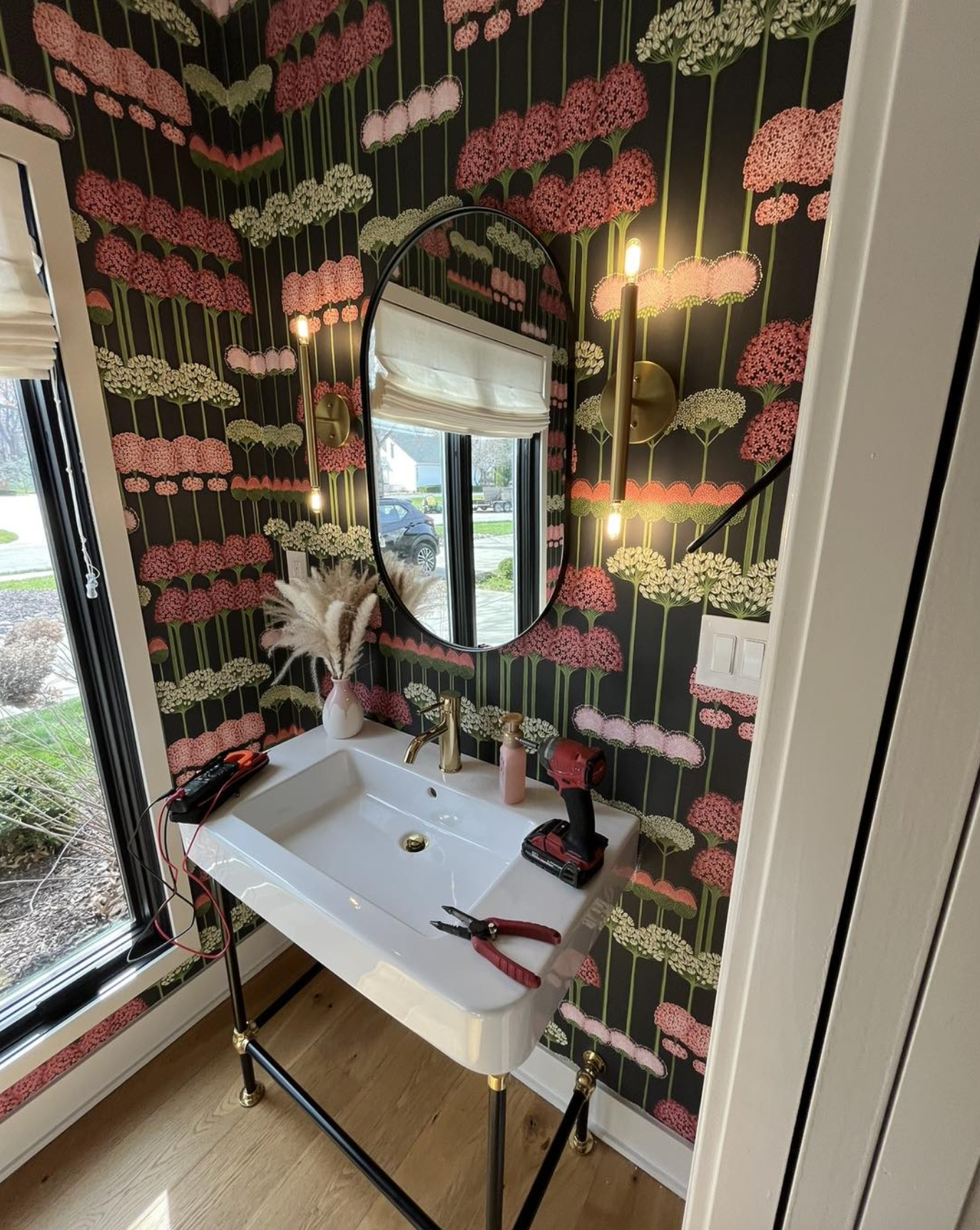 Bathroom with black wallpaper featuring pink and white flowers, oval mirror, white sink, golden faucet, and light fixtures. Tools, a hairdryer, and a vase with pampas grass are on the sink.