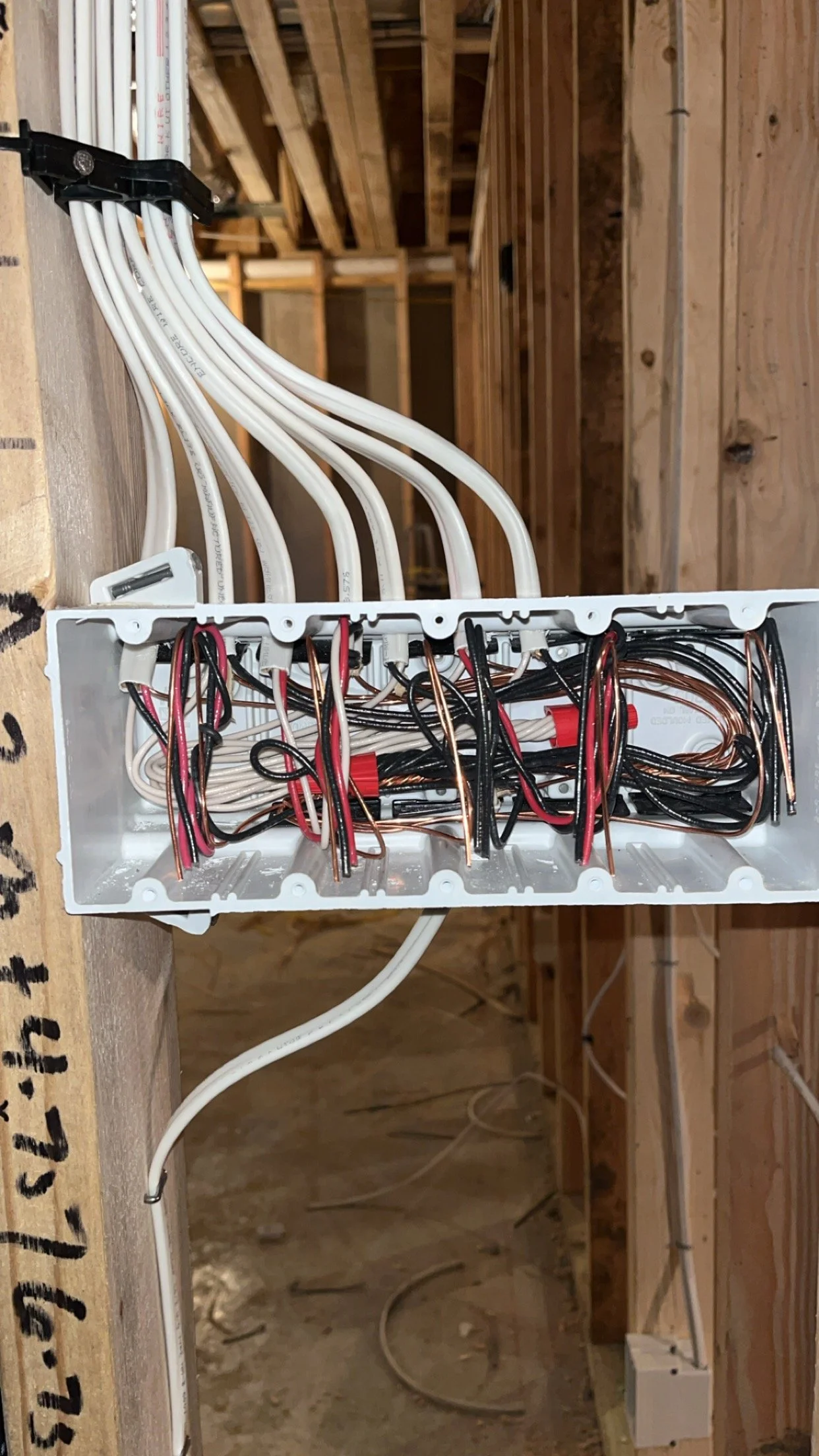 An electrical wiring box with white, black, red, and copper wires connected, mounted on wooden studs inside a building under construction.
