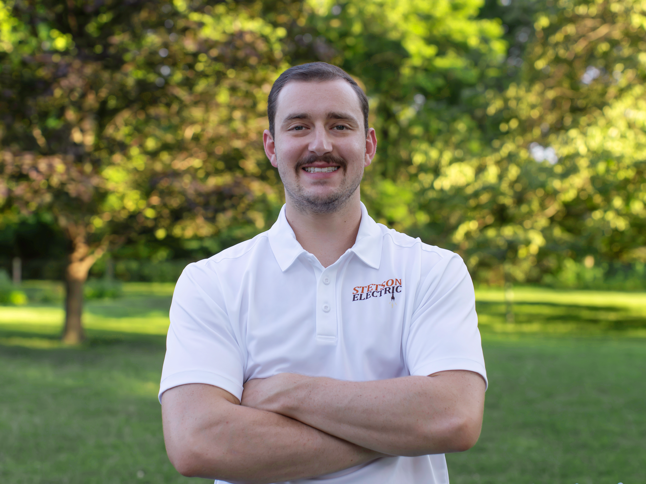 A young man with short dark hair, light skin, and a goatee, smiling with arms crossed, standing outdoors in front of trees on a sunny day, wearing a white shirt with a logo that reads 'Stetson Electric'.
