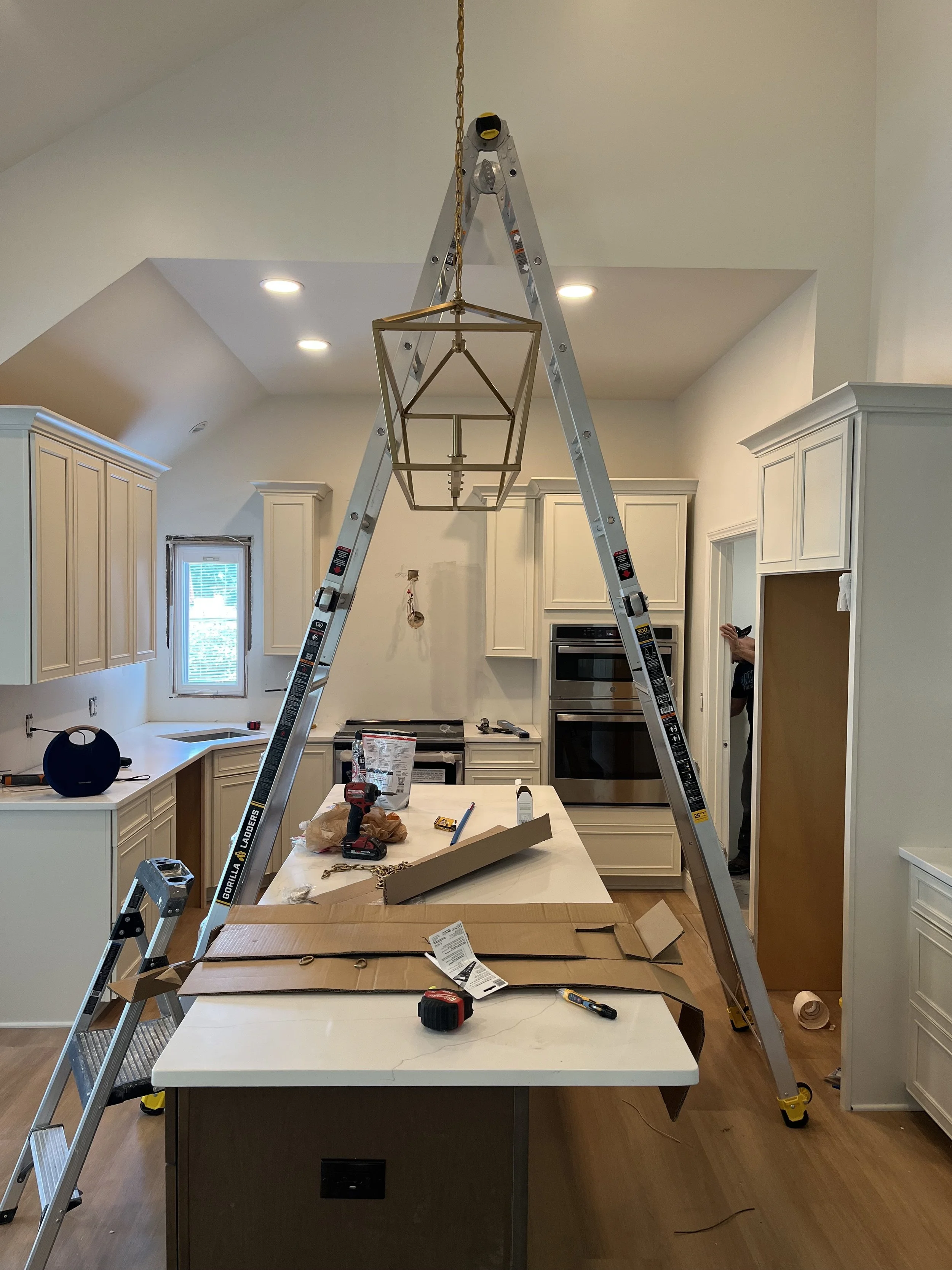 Kitchen with white cabinets and a large island. A step ladder is set up in the middle, hanging a light fixture. Tools, boxes, and materials are on the island and around the kitchen, indicating ongoing installation or renovation work.