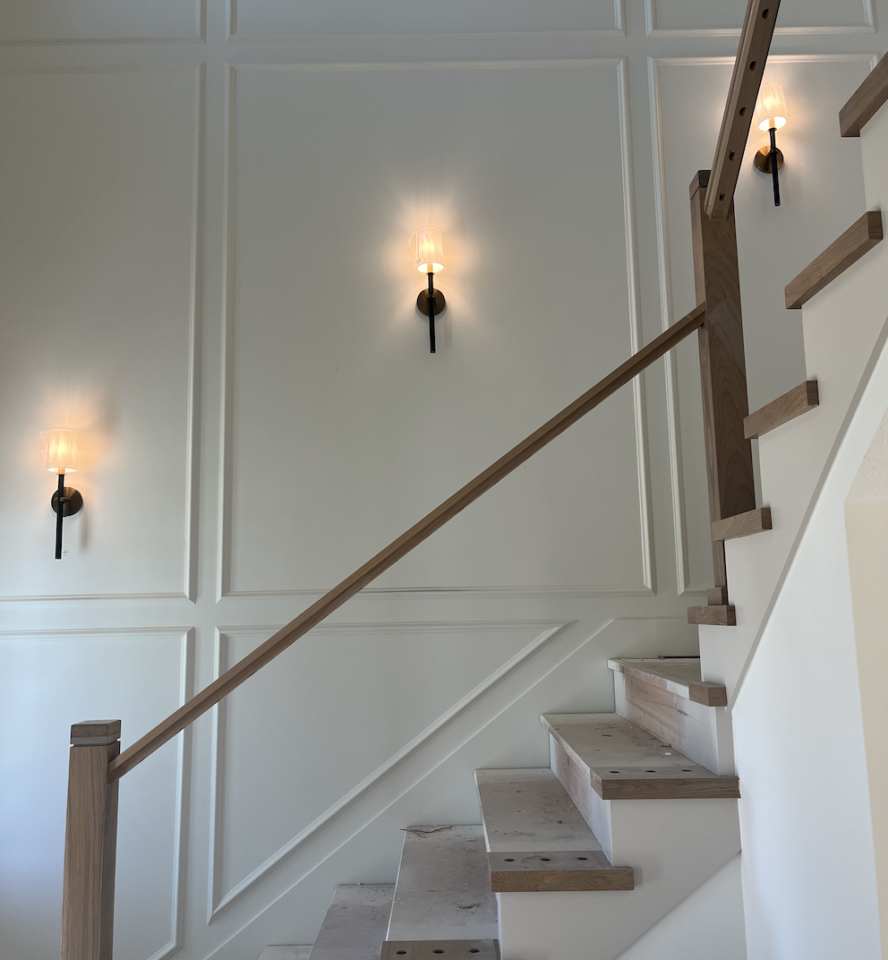 Interior view of a staircase with unfinished steps, a wooden handrail, and three wall sconces on a white paneled wall.