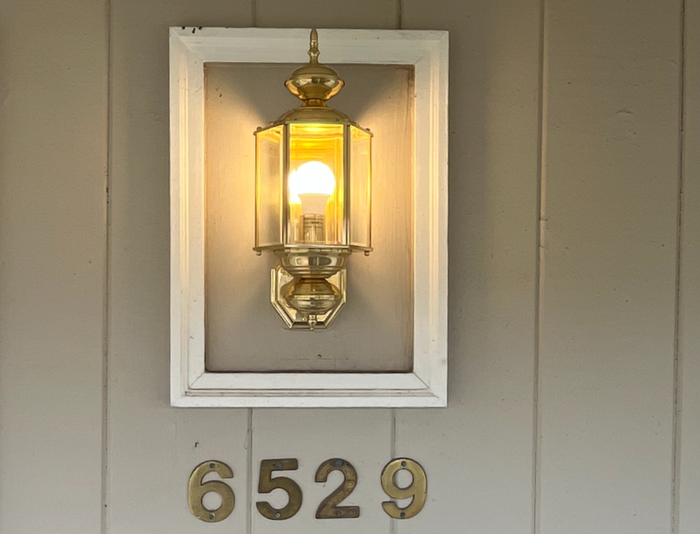 A wall-mounted lantern-style light fixture with a visible bulb, illuminating the area. Below the light, house numbers "6529" are displayed in gold-colored metal plaques.