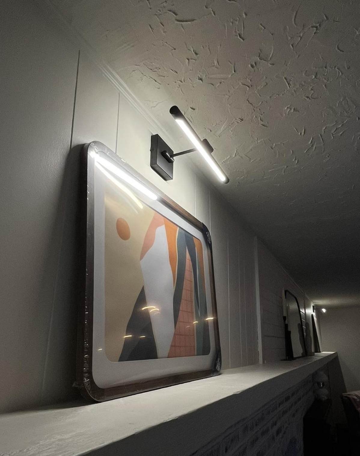 Close-up of a picture frame with modern abstract art on a white shelf, illuminated by a ceiling-mounted LED light fixture in a room with textured ceilings and neutral-colored walls.