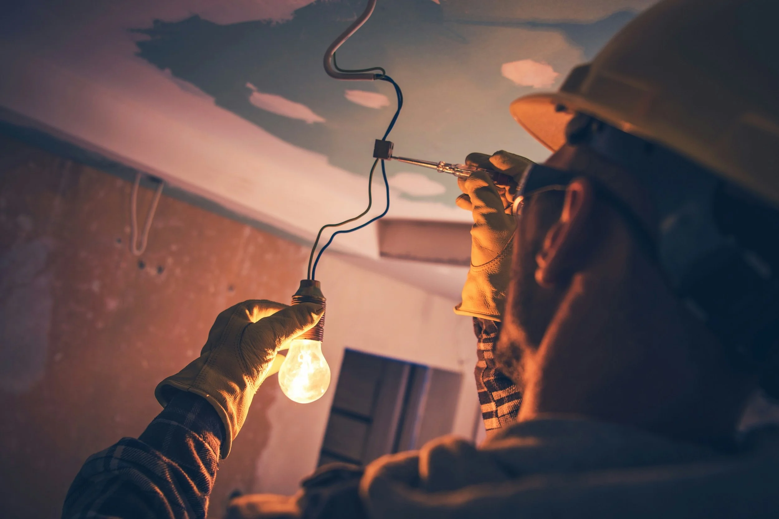 A person in gloves holding a light bulb, while another person uses a screwdriver on an electrical wire in the ceiling.