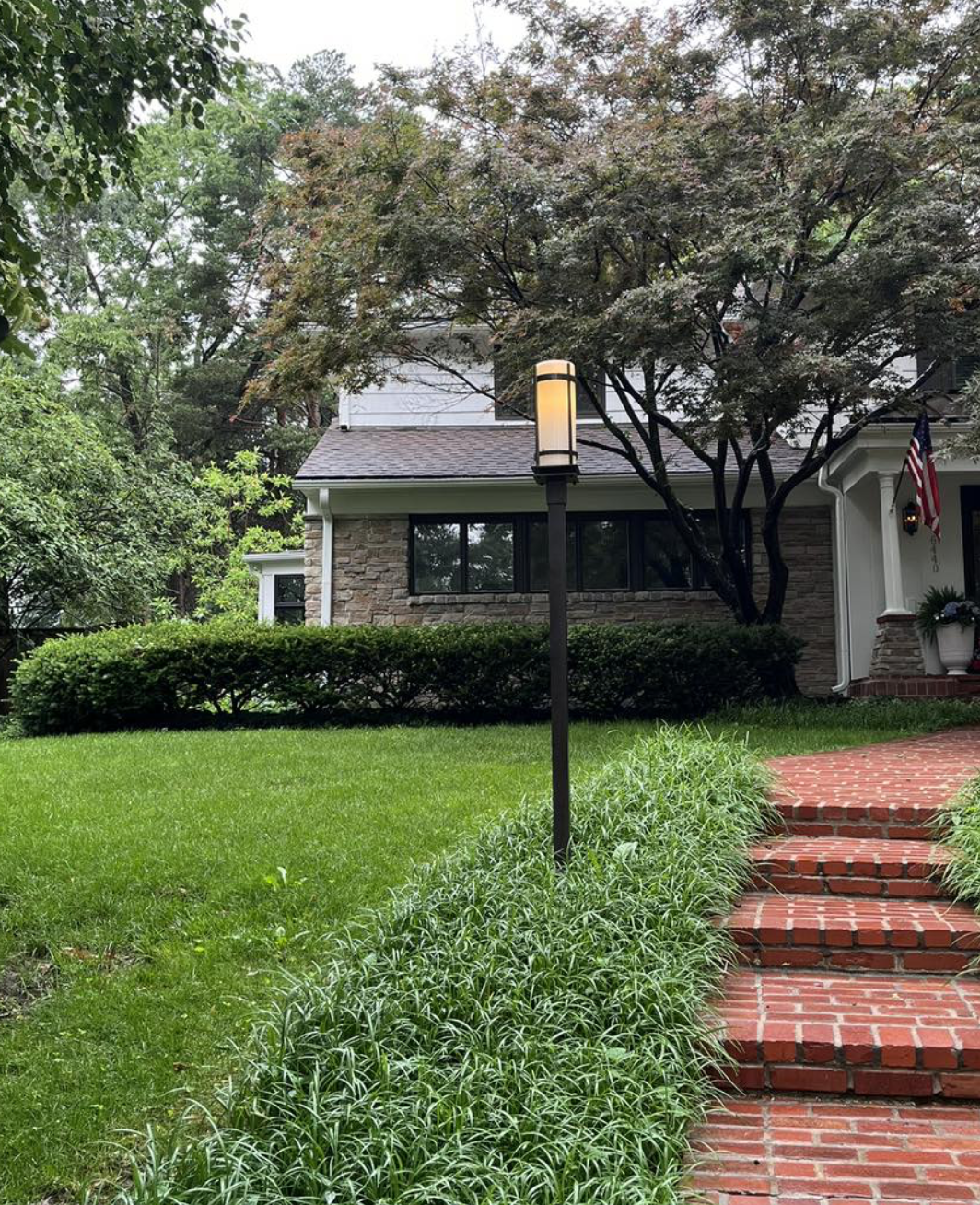 A brick pathway with steps leading to the front porch of a house surrounded by green grass, bushes, and trees. A street lamp is standing along the path, and an American flag is displayed near the house.