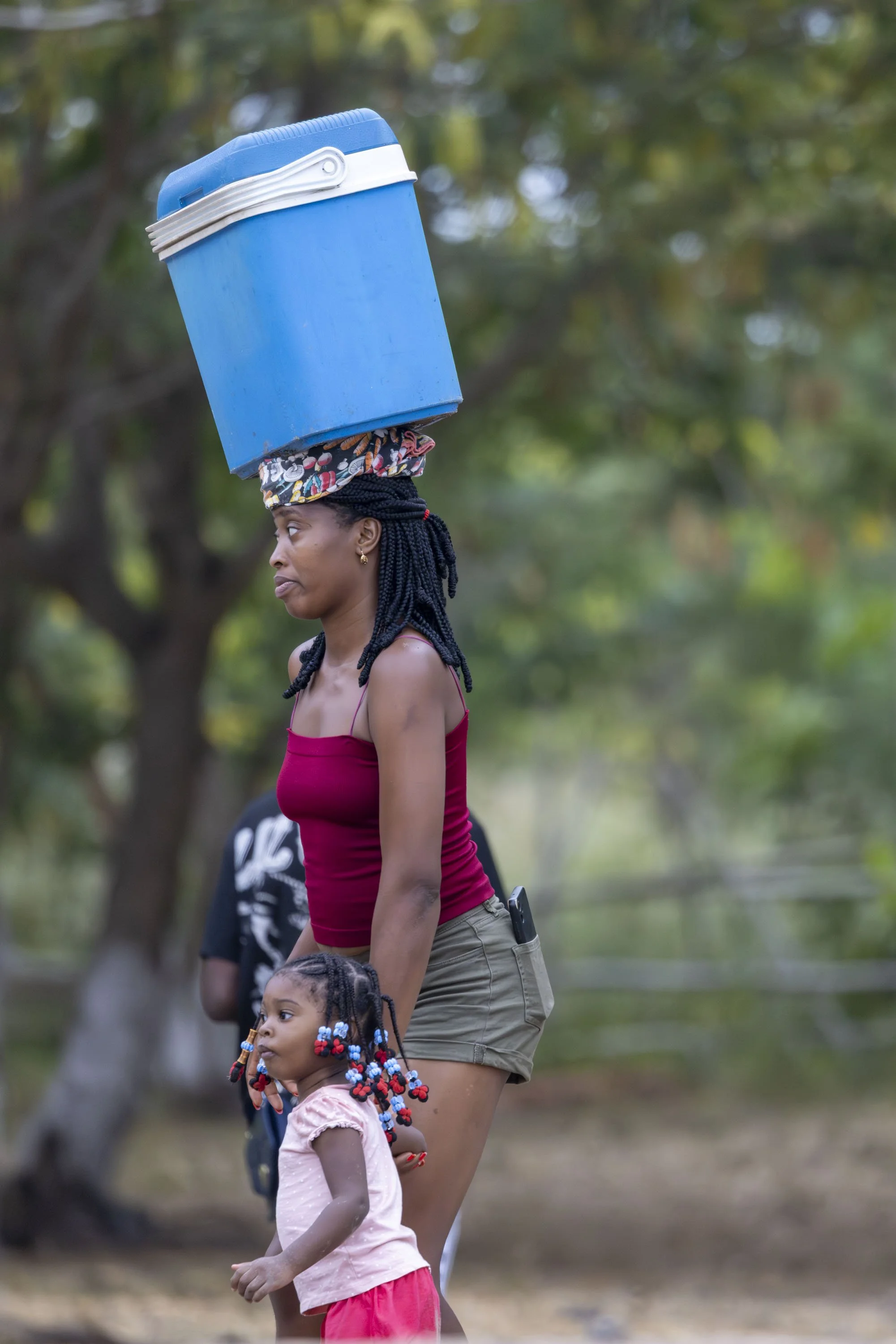 A woman with braided hair and a red tank top balancing a blue cooler on her head while holding a young girl with colorful braids in an outdoor park with trees in the background.