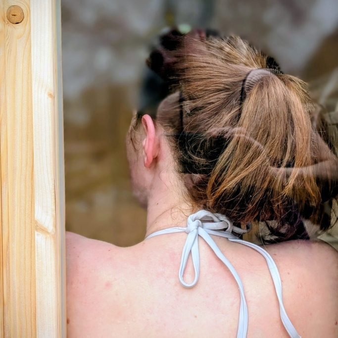 A woman with her hair in a messy bun, seen from behind, standing next to a wooden structure outdoors.