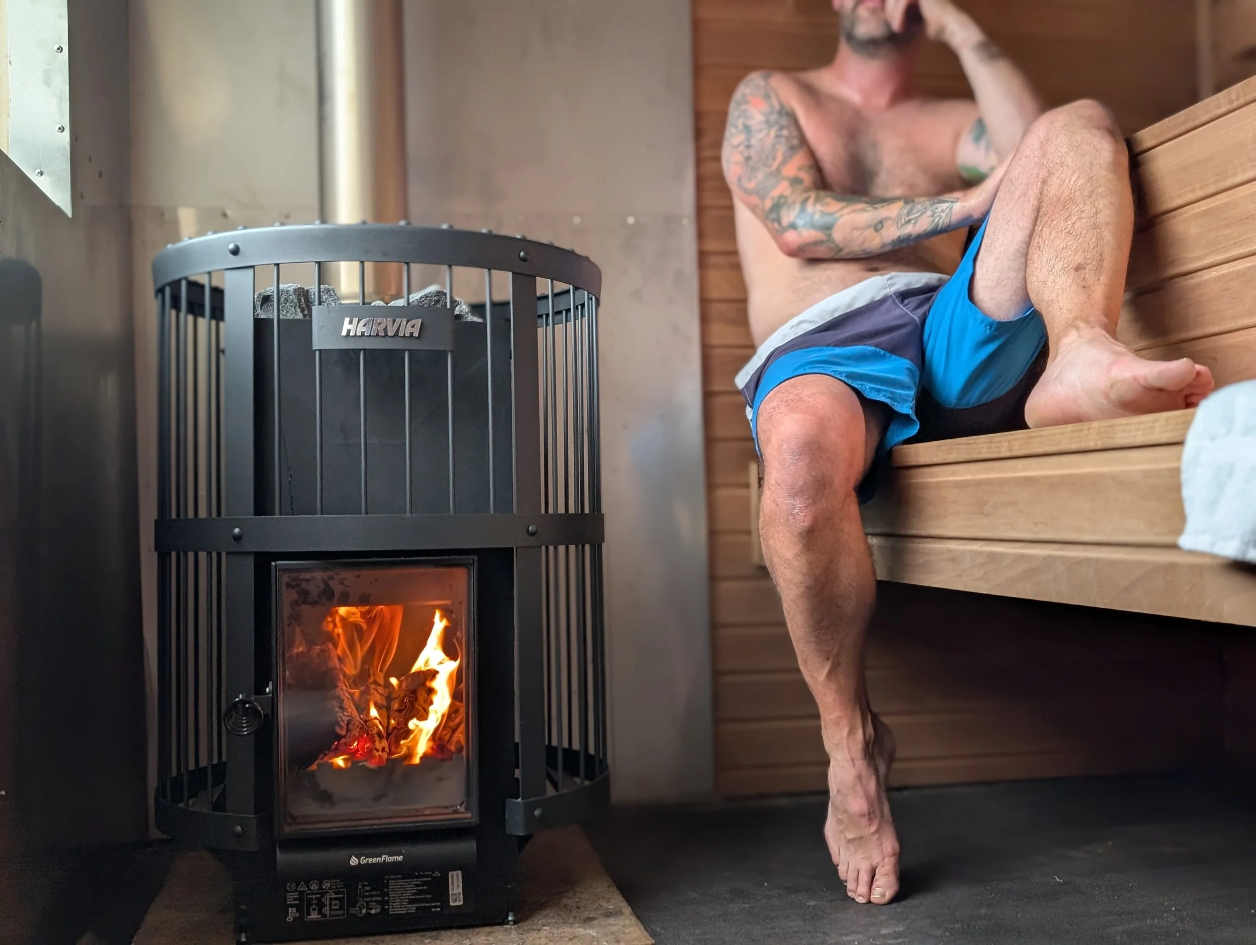 A shirtless man with tattoos sitting on a wooden bench in a sauna, looking away with one hand resting on his head, next to a black wood-burning stove with flames visible.