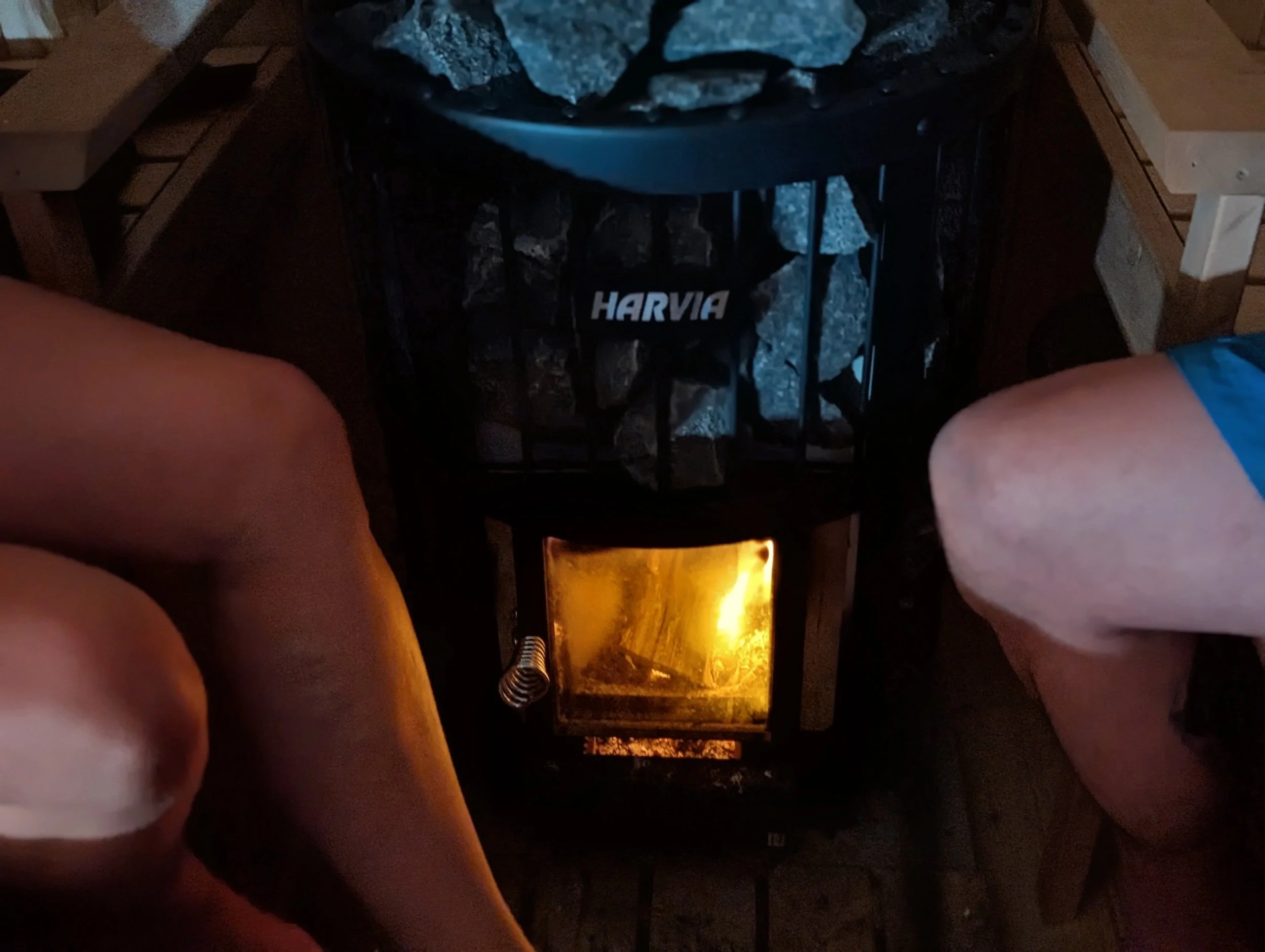 A person's legs are visible on each side of a woodstove with a glass door showing the fire inside. The person is wearing shorts, and the surrounding area appears to be a cozy indoor setting.